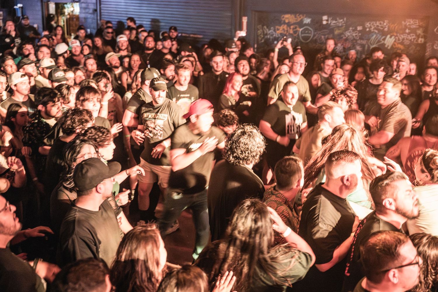A densely packed concert crowd fills a dimly lit venue with stage lighting casting an orange glow over the audience.
