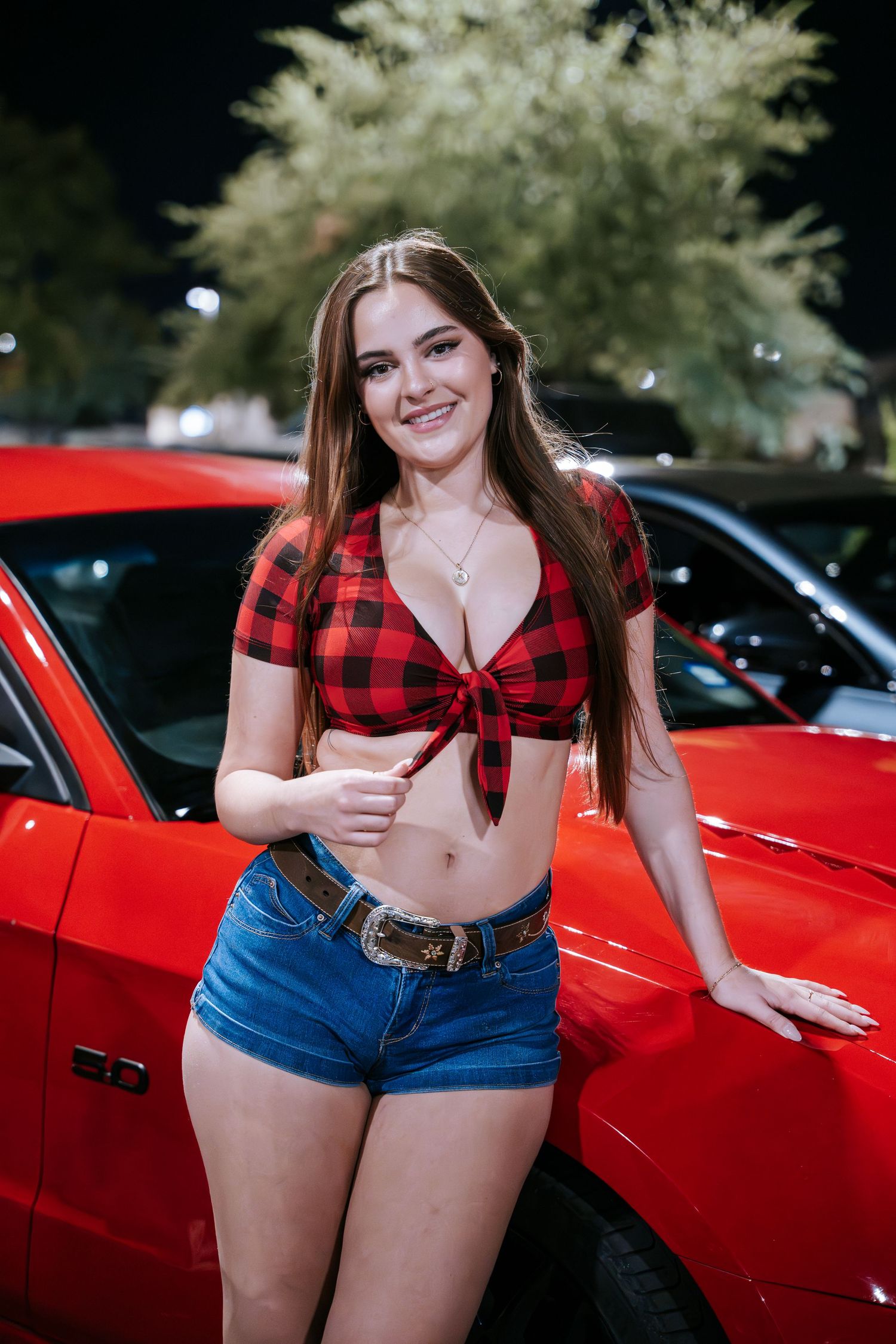 Model in red plaid crop top and denim shorts poses next to a bright red car at night.