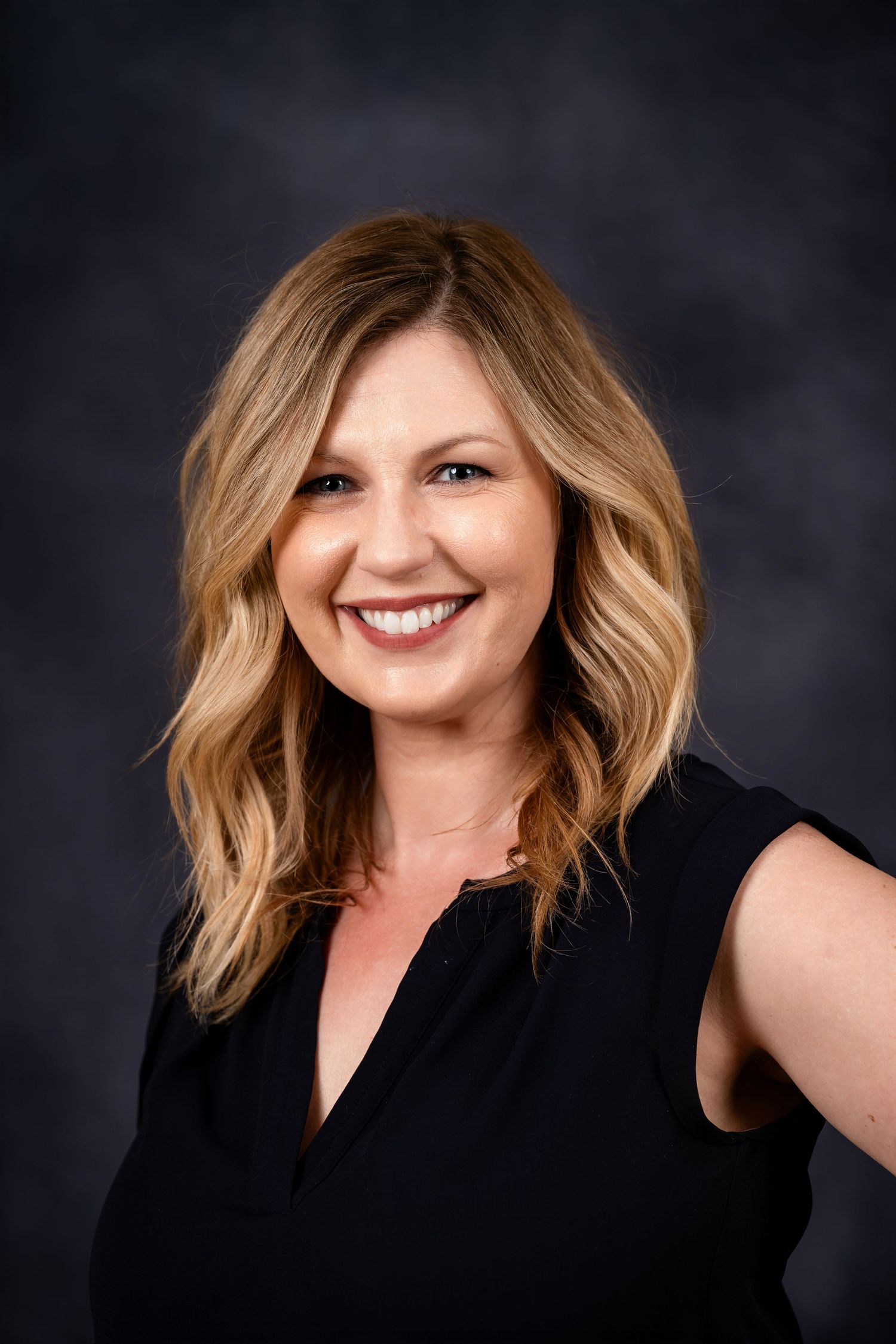Studio headshot featuring wavy blonde hair and black sleeveless top against dark background.