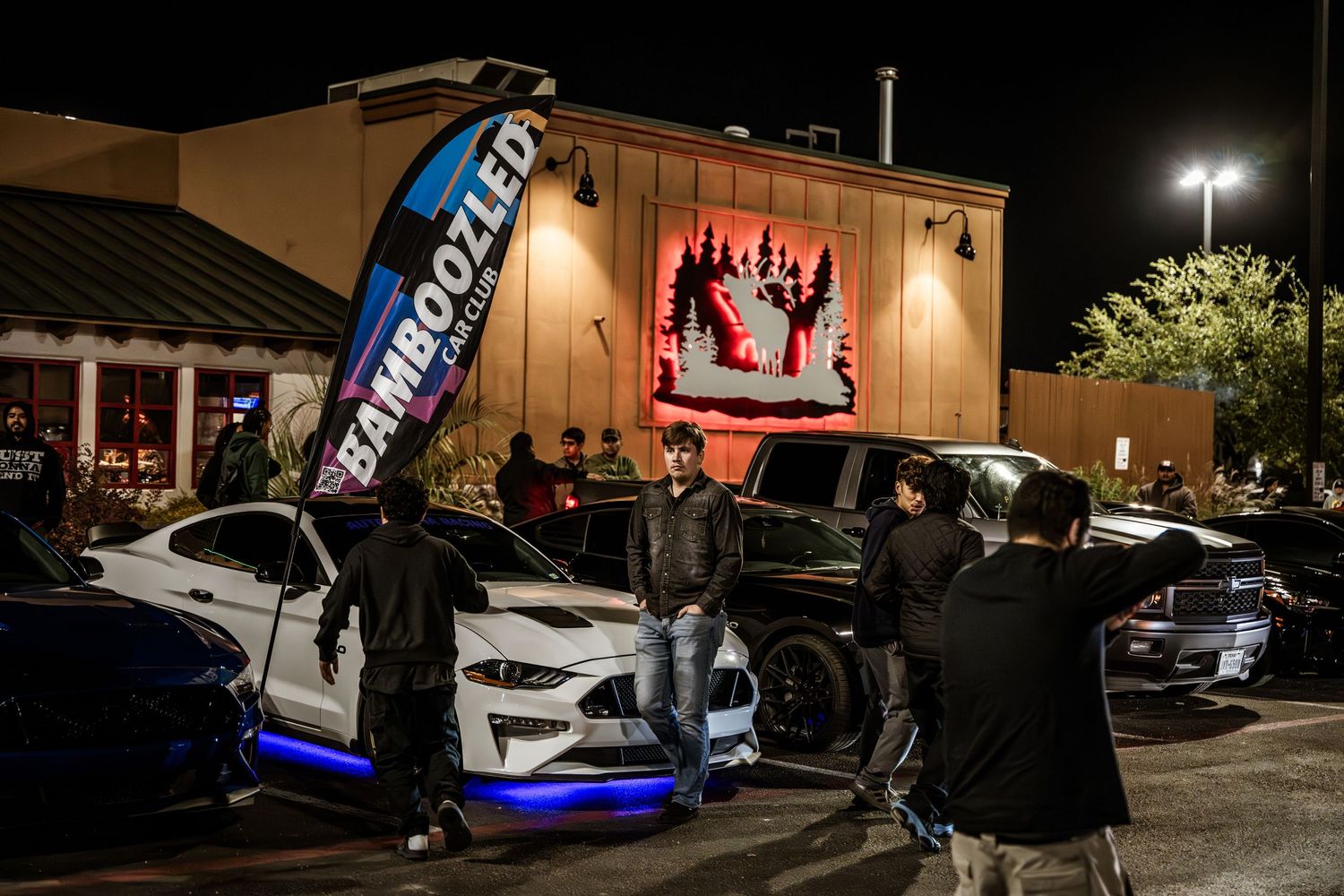 Nighttime car meet showing modified vehicles with LED lights and large display screen.