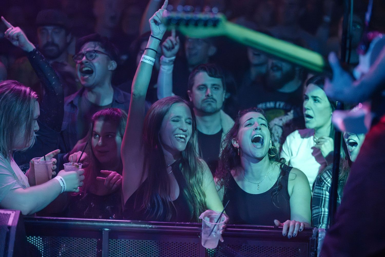Enthusiastic concert crowd cheers and raises hands under colorful venue lighting.