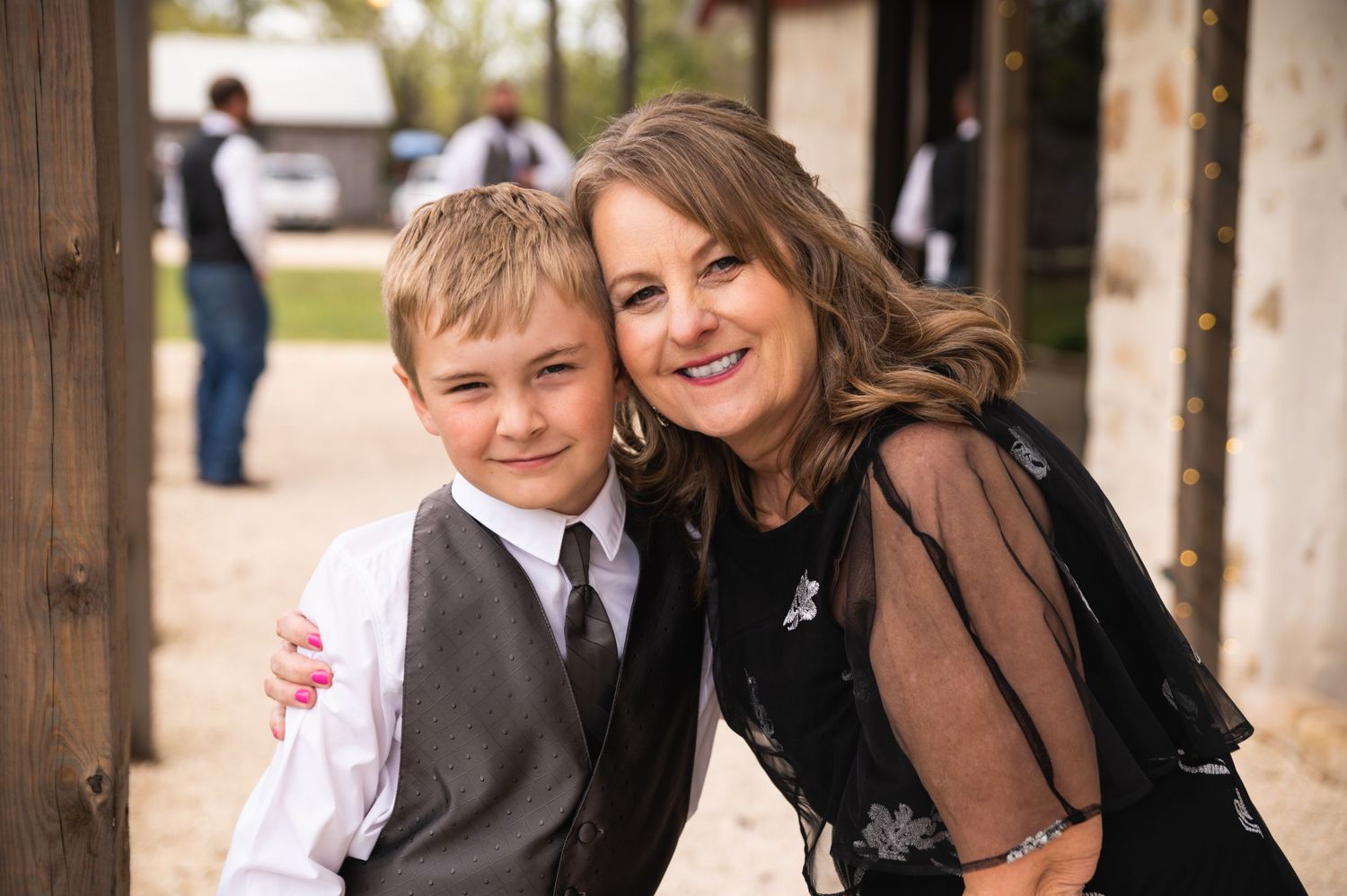 A young boy in formal attire and someone wearing a brown sheer top pose together at an outdoor event.