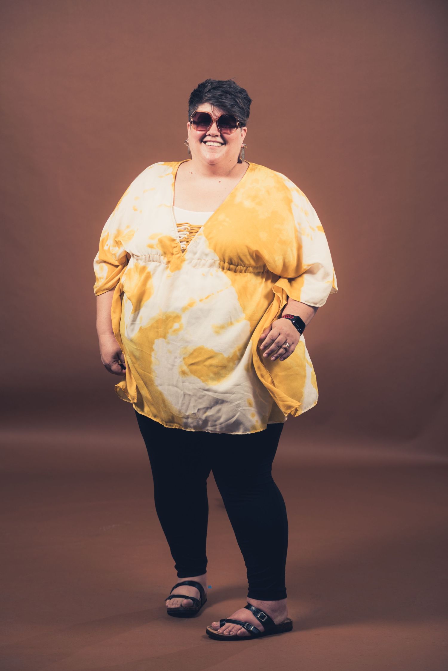 Full-length portrait of person wearing yellow tie-dye outfit and sunglasses against brown background.
