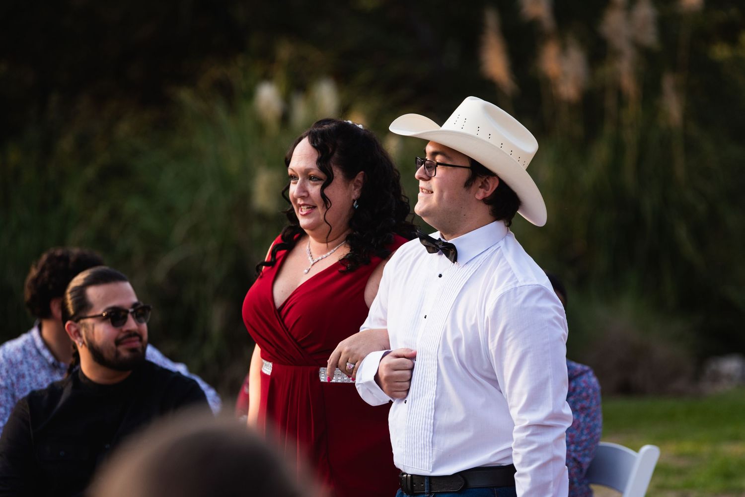 Wedding guests enjoy festivities outdoors with person wearing cowboy hat and formal wear.
