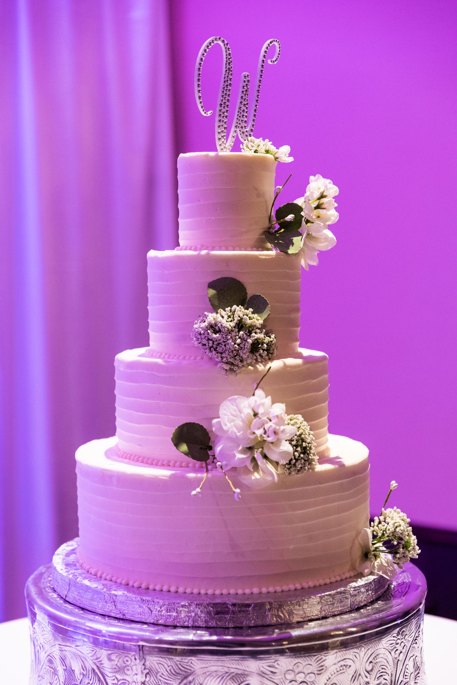 Four-tier white wedding cake with crystal monogram topper and floral decorations against purple backdrop.