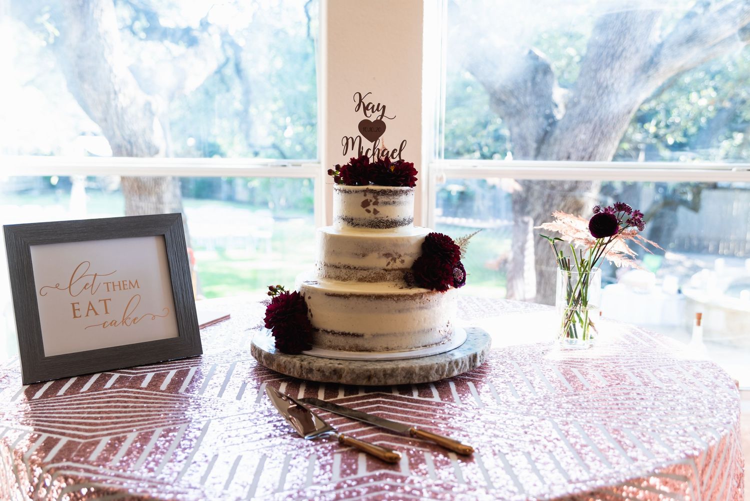 A semi-naked white wedding cake decorated with burgundy flowers sits on a lace tablecloth.