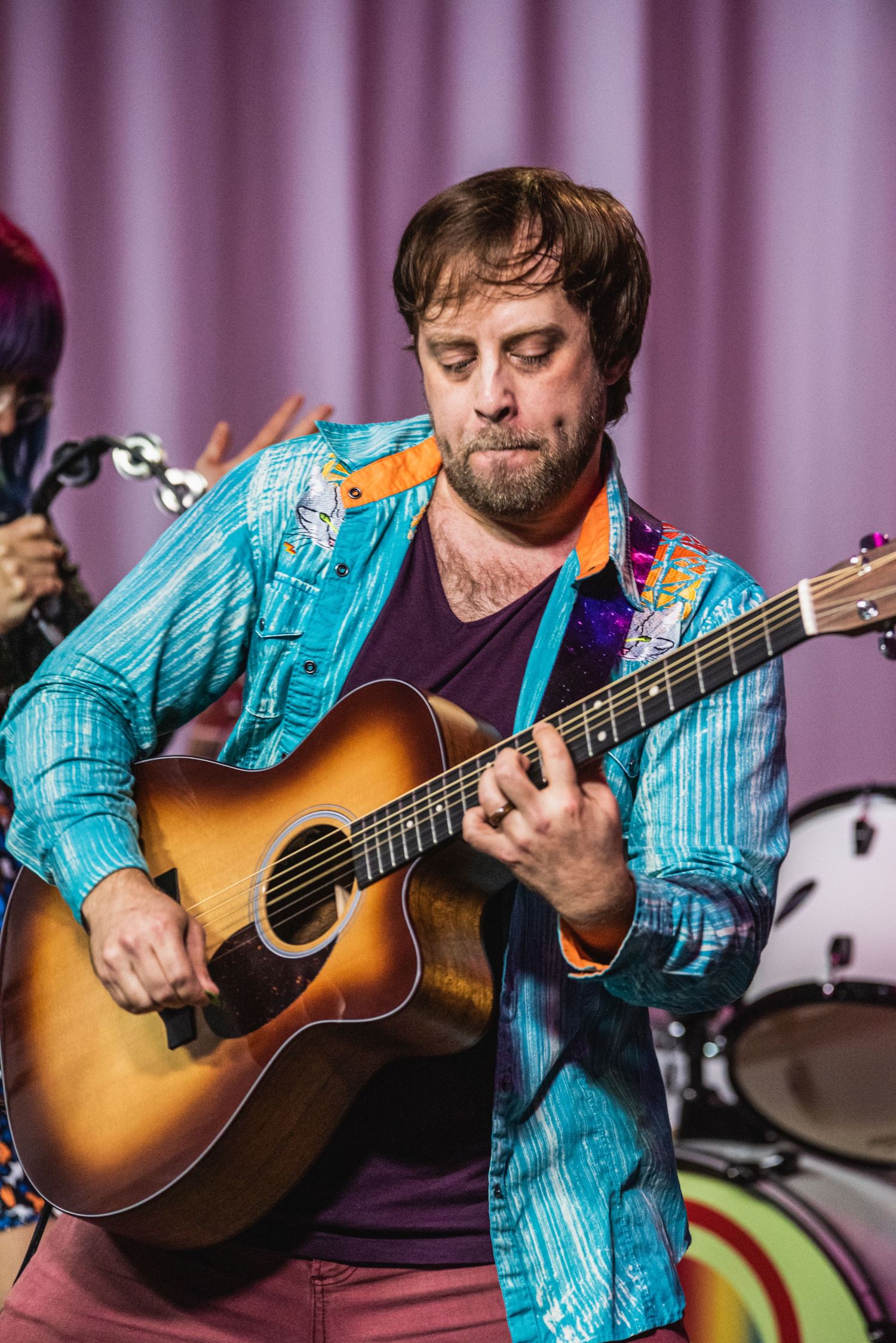 Performer in patterned turquoise jacket concentrates while playing acoustic guitar on stage.
