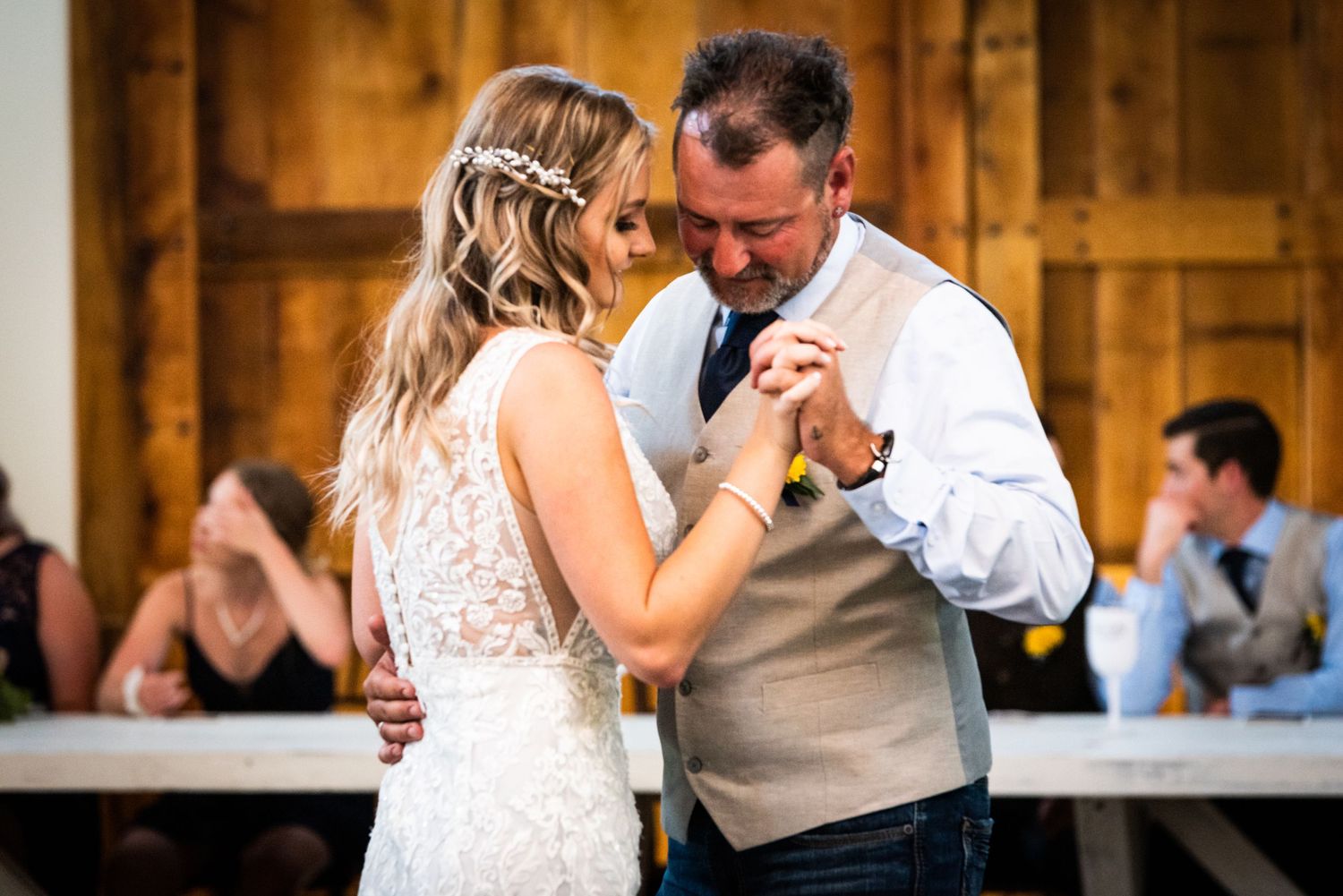Tender moment during wedding dance in rustic barn venue with guests in background.