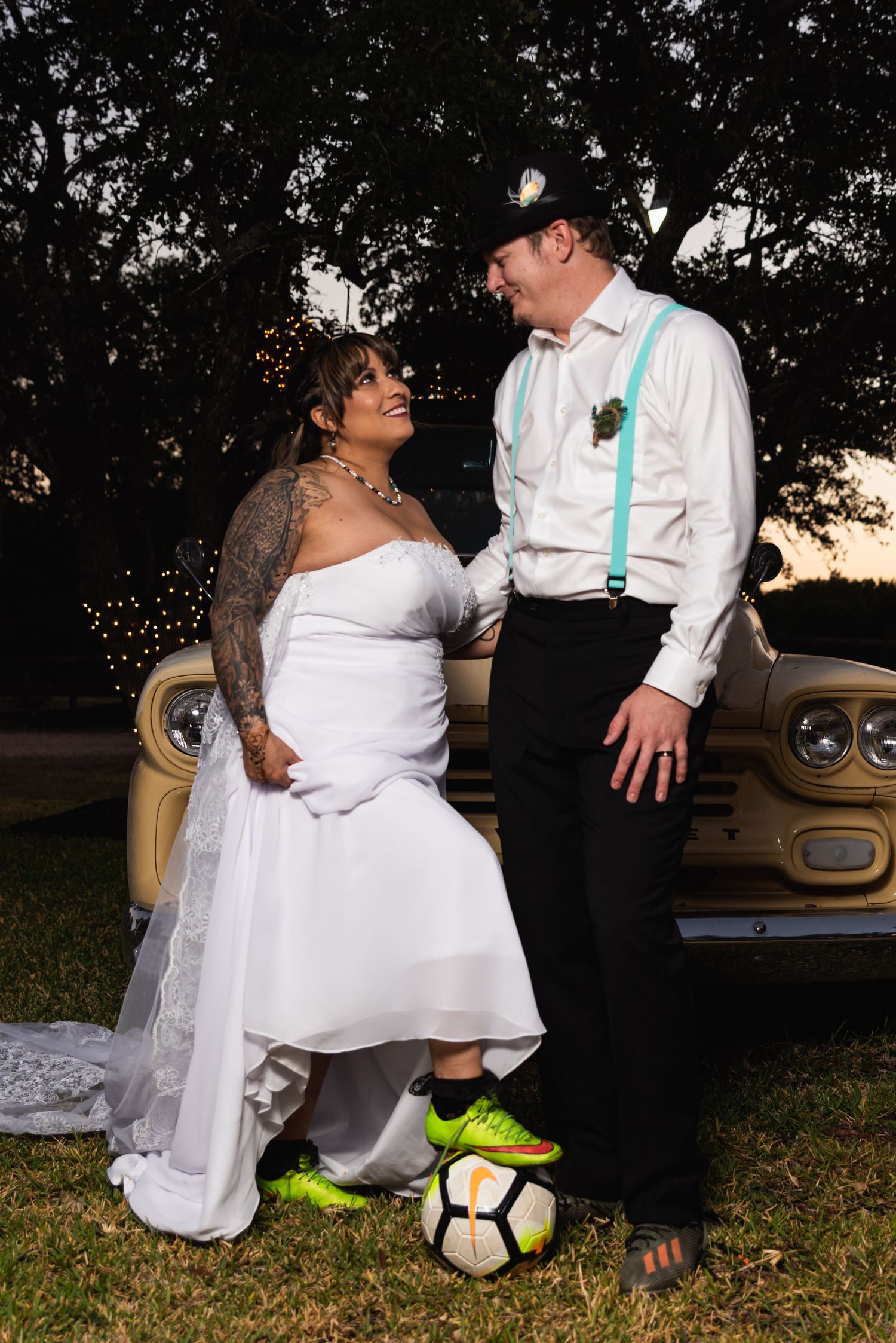A couple in wedding attire poses with a soccer ball near a vintage car at sunset.