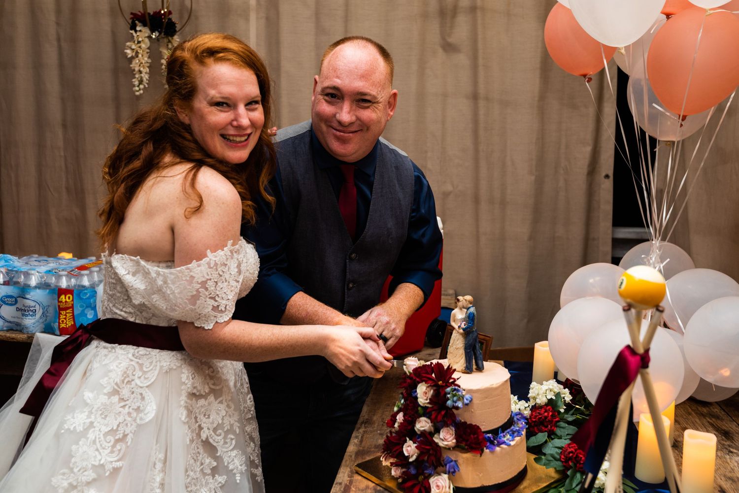 A couple cuts their wedding cake together at their reception surrounded by balloons and decorations.