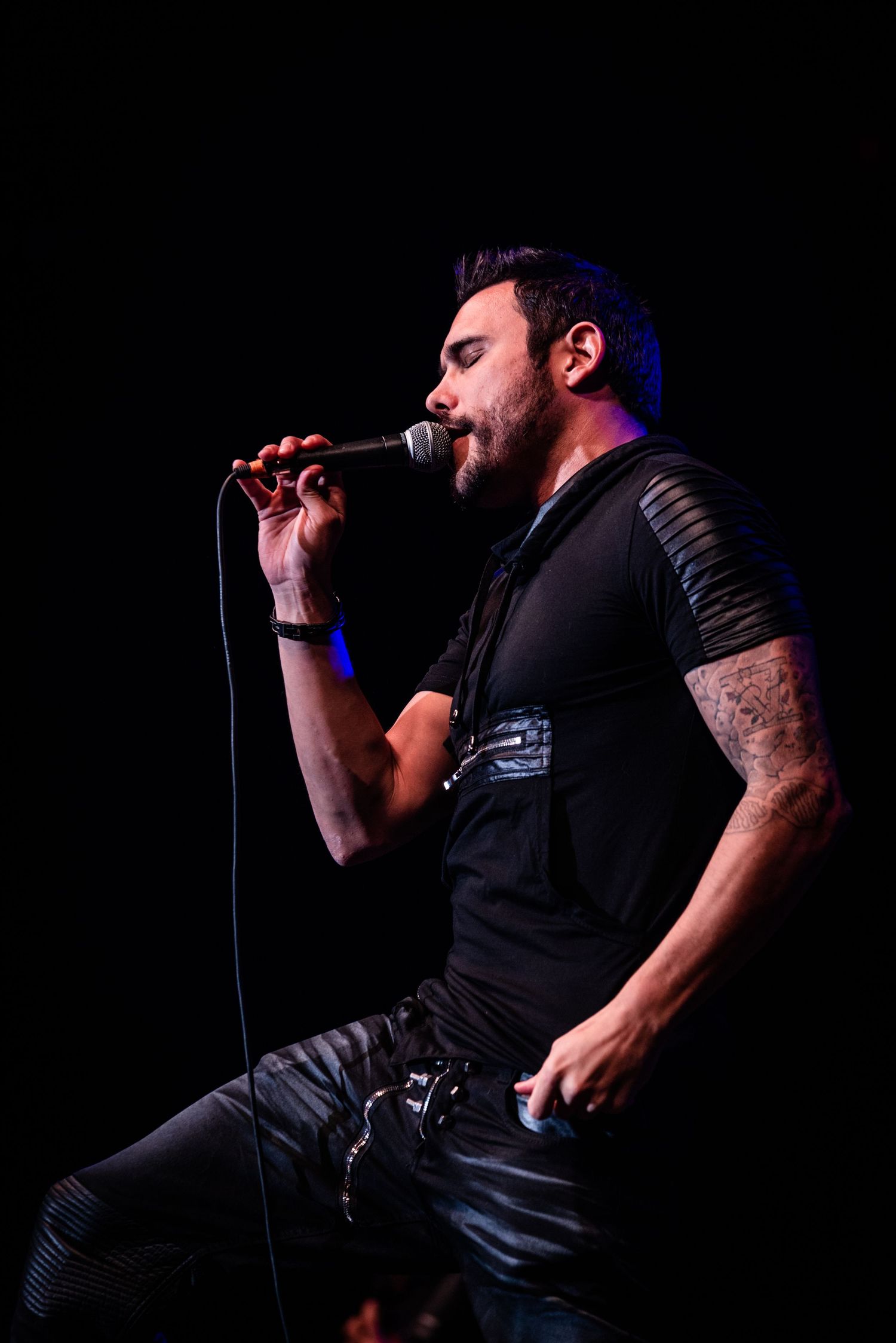 Vocalist in black attire performs energetically on stage with dramatic lighting and microphone.