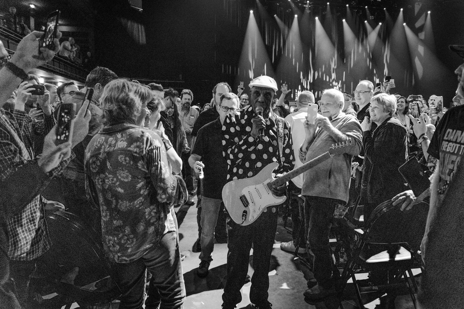 Concert scene showing performer in polka dot shirt surrounded by audience members in black and white.