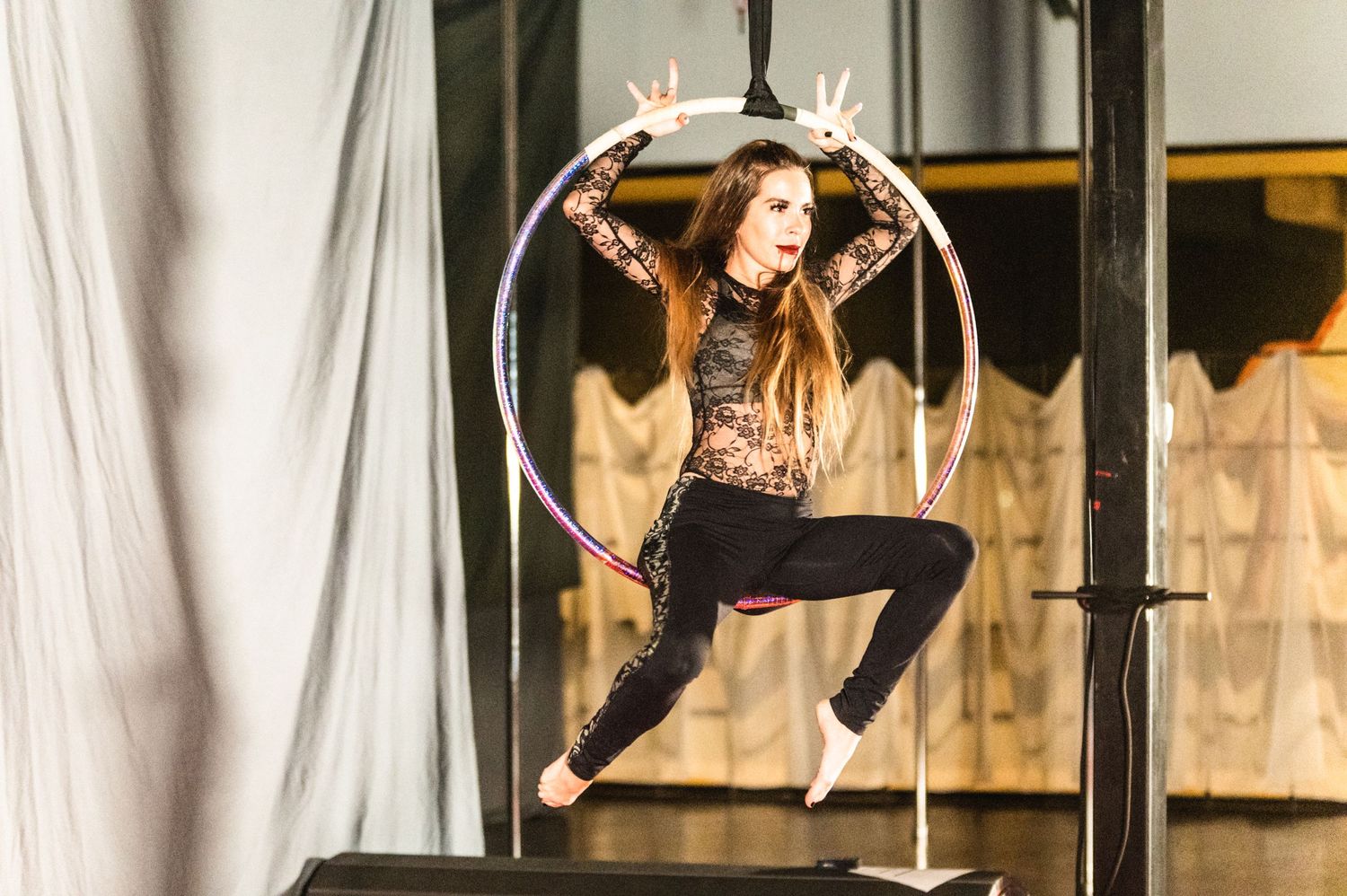 Acrobat performs on aerial hoop with dramatic lighting and white curtain backdrop.