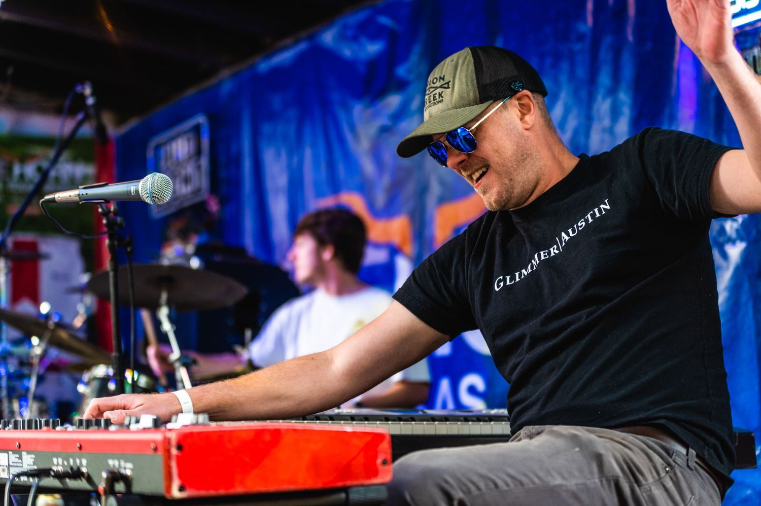 Musician plays red keyboard while wearing black t-shirt and baseball cap at outdoor venue.