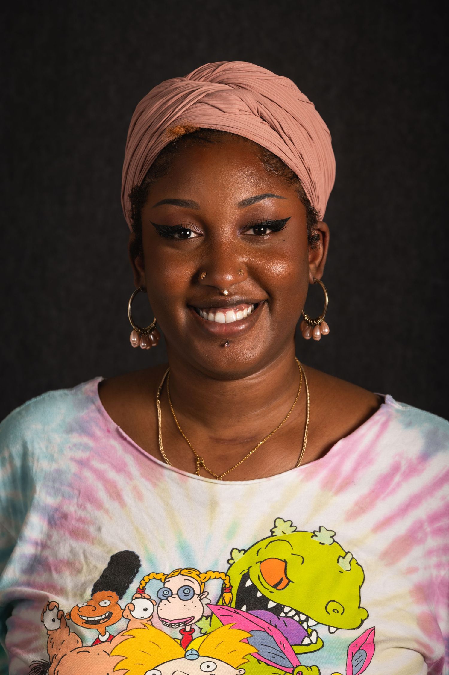 Portrait of a person in tie-dye Rugrats shirt wearing a peach headscarf and jewelry.