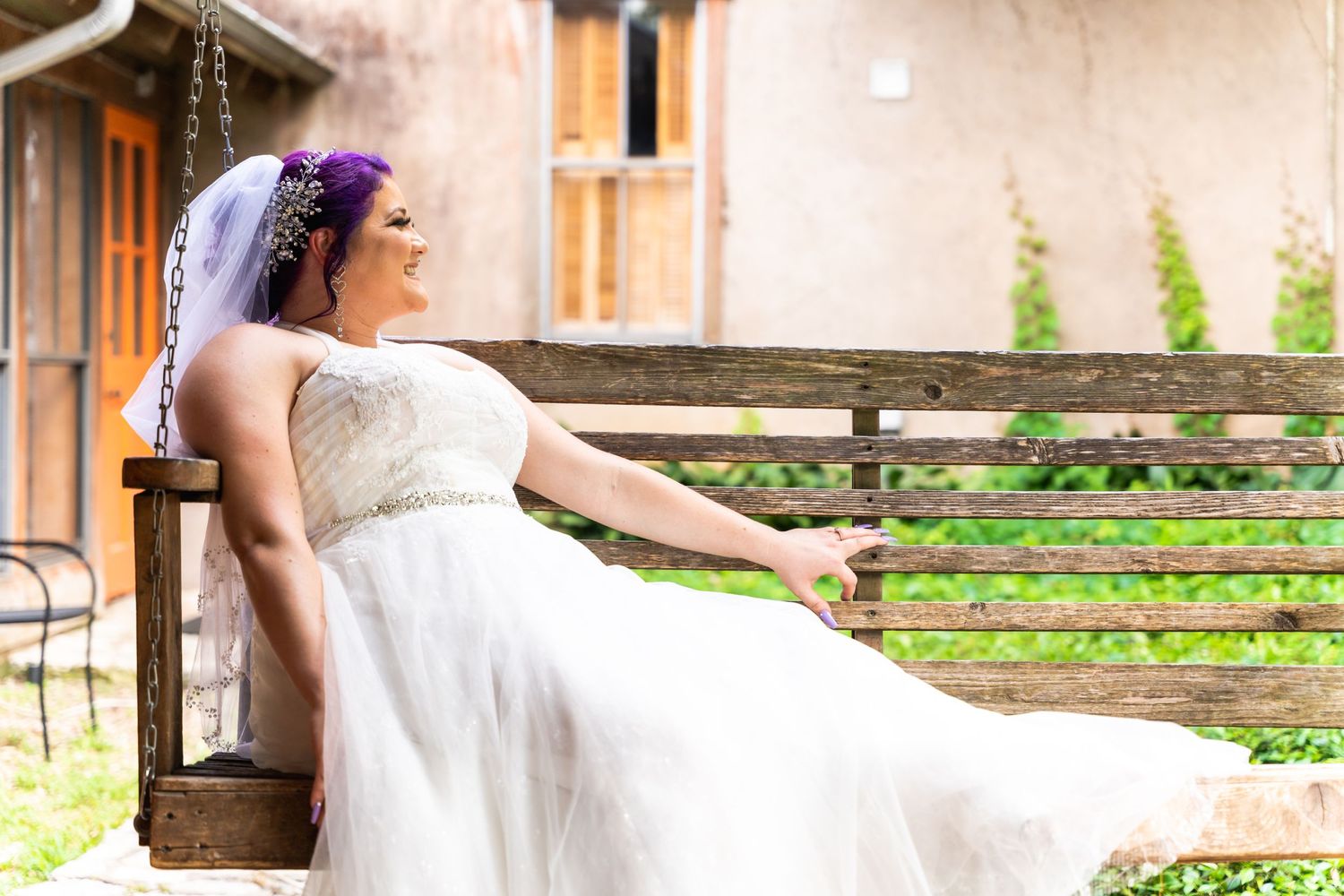 A bride in a white wedding dress with purple hair rests on a rustic wooden bench.