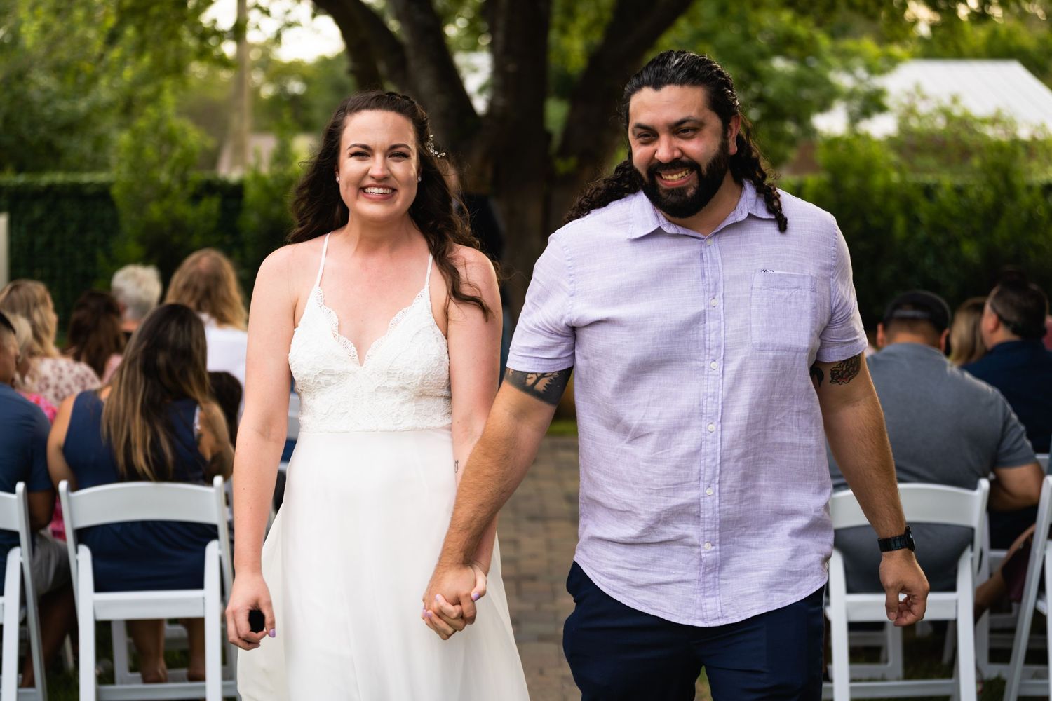 A couple walks together outdoors at a wedding reception with chairs visible in background.