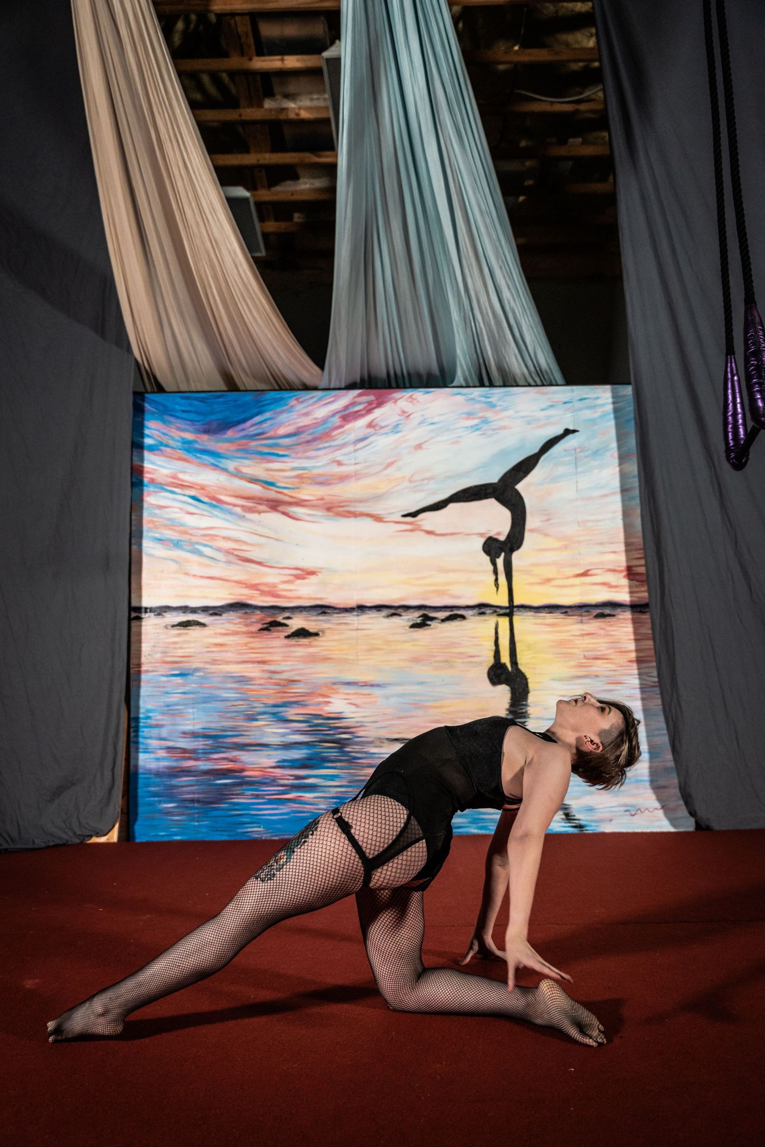 Dancer in black outfit poses dramatically against colorful sunset backdrop with aerial silks.