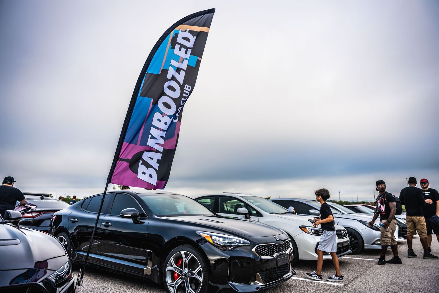 Car meet gathering with BMW vehicles under Eurooozer feather flag banner.