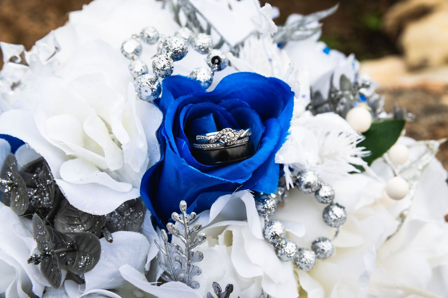 An engagement ring sits in a blue rose surrounded by white flowers and silver accents.