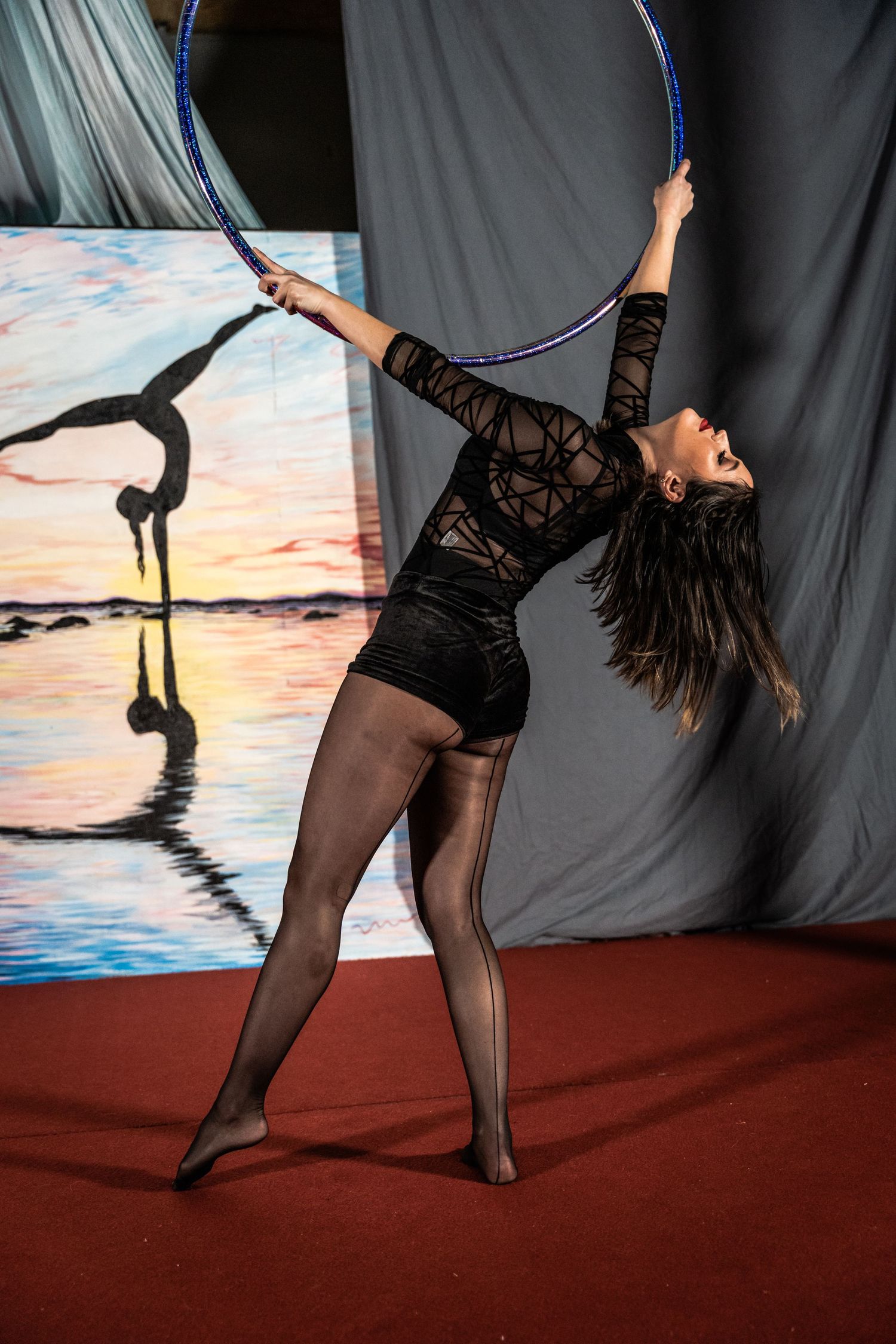 Performer in black leotard and tights gracefully posing with a hoop against painted backdrop.