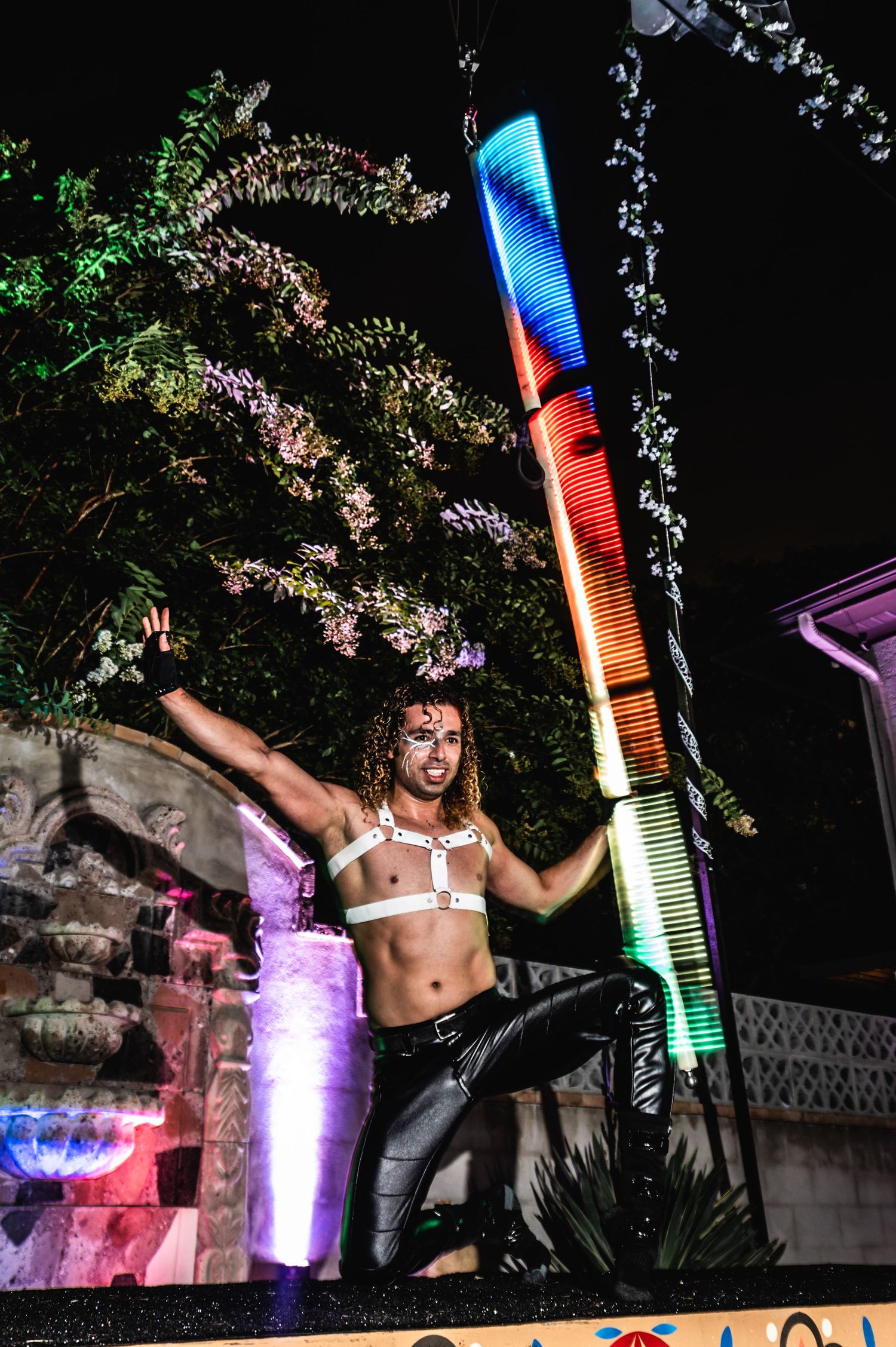 Artist performs dramatic pose next to tall multicolored light installation at night.