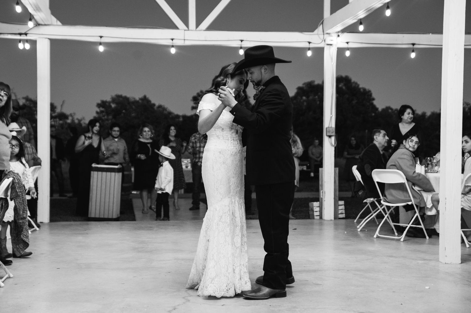 Black and white photo of newlyweds sharing first dance under string lights in an outdoor reception venue.