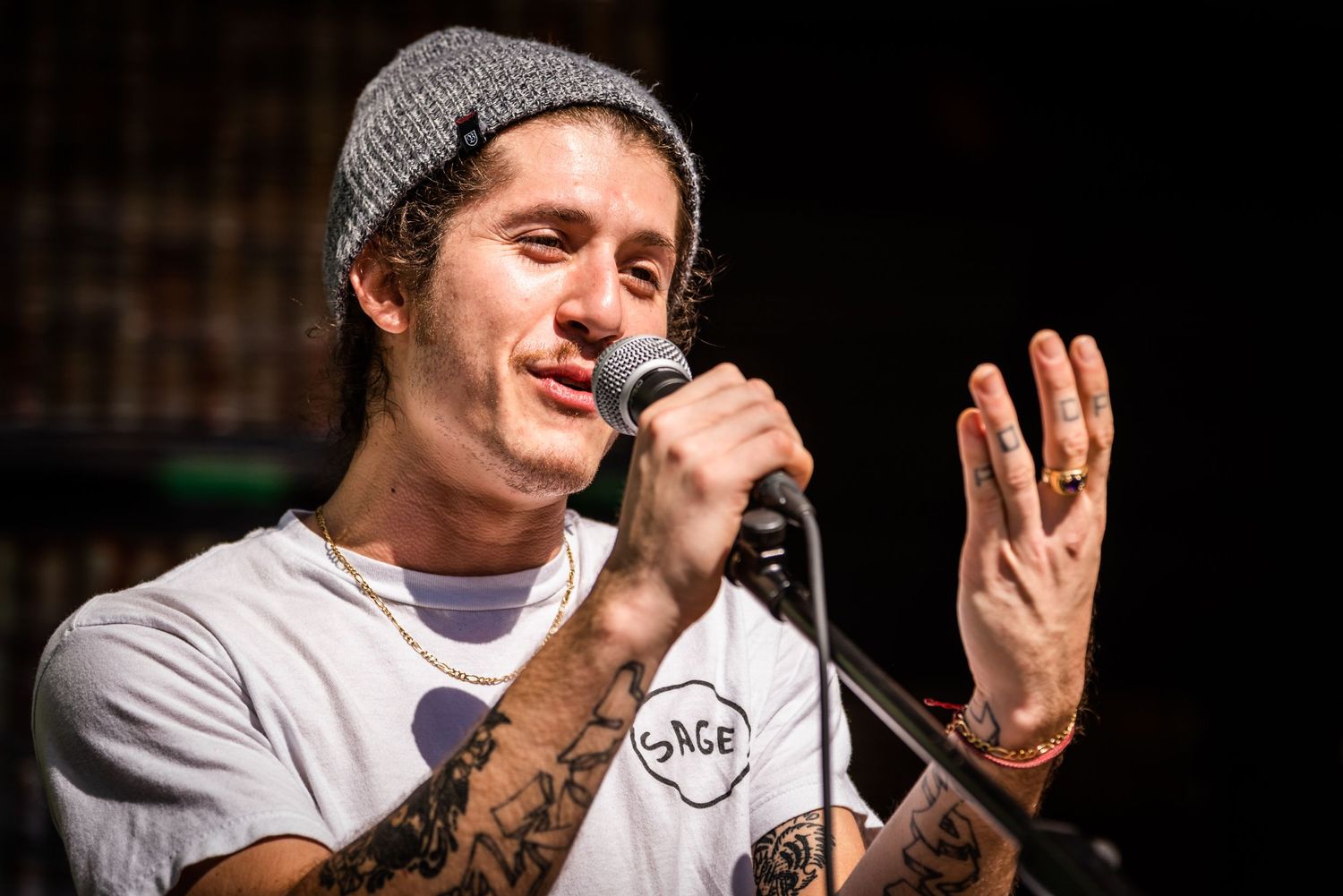 A performer with a grey beanie sings into a microphone while gesturing on stage.