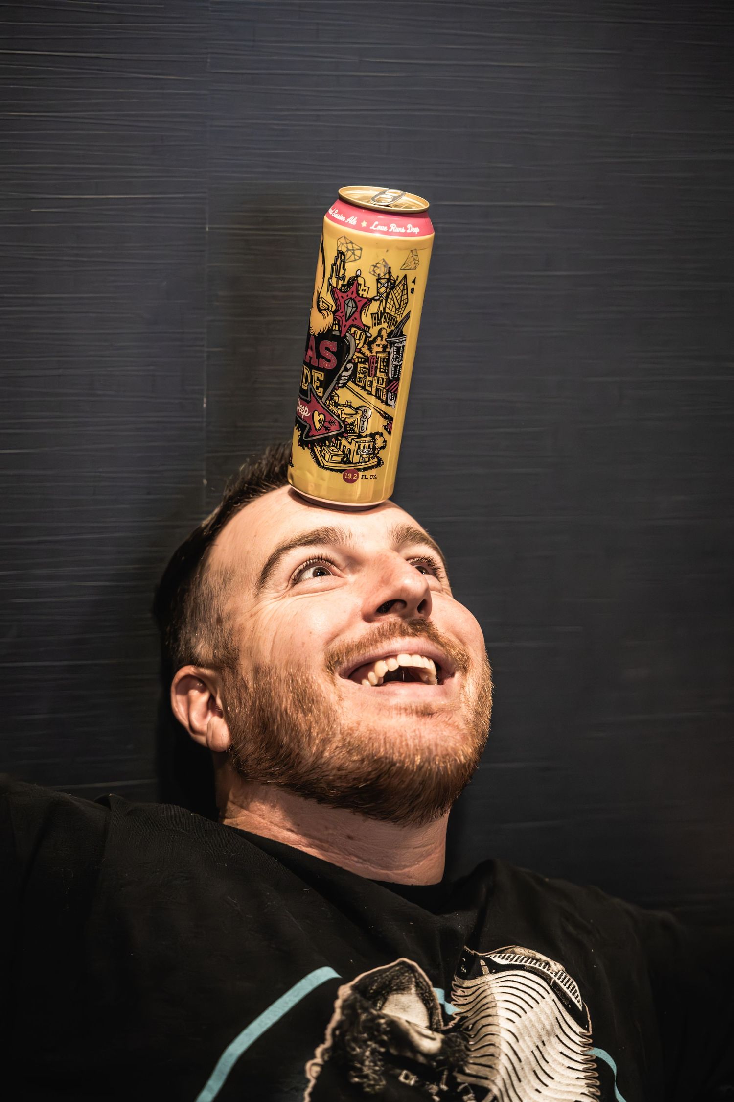 Playful portrait balancing energy drink can on forehead against black background.