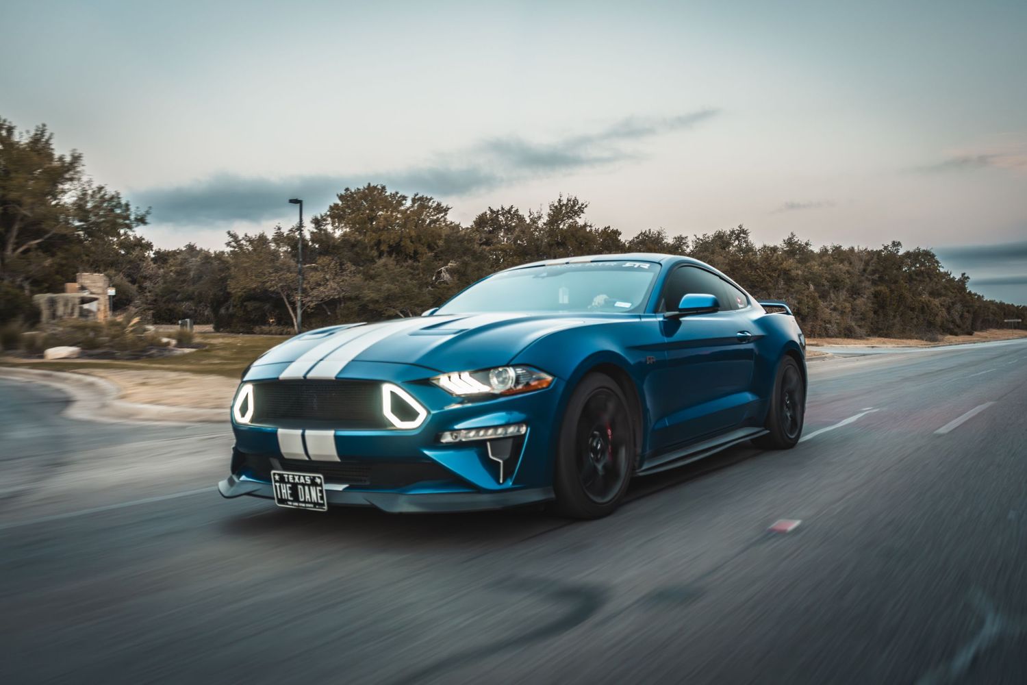 Blue Ford Mustang GT with racing stripes speeding on open road.
