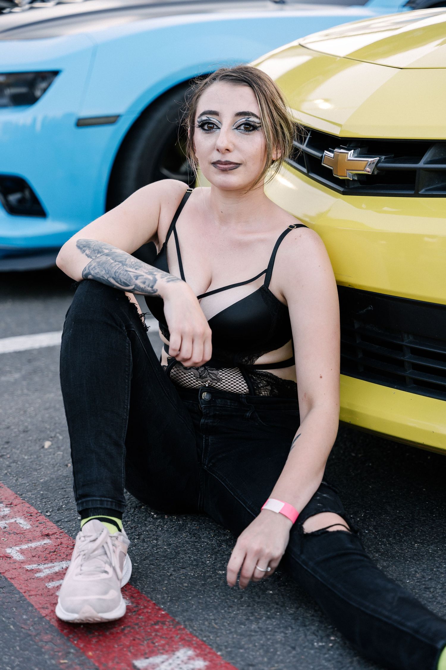 A person in black attire sitting next to a yellow Chevrolet Camaro at a car show.