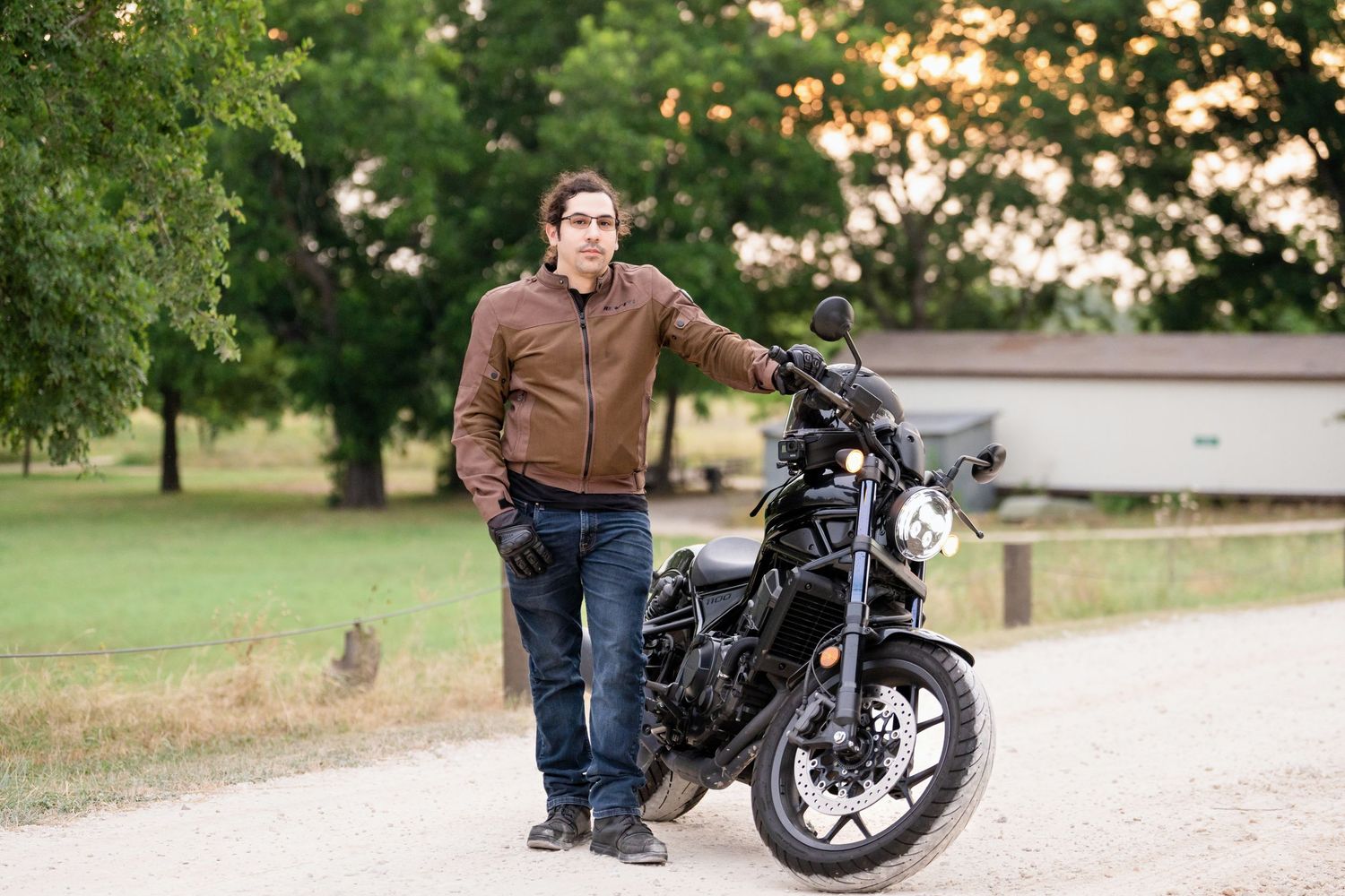 A person stands confidently next to a sleek black motorcycle on a park trail.