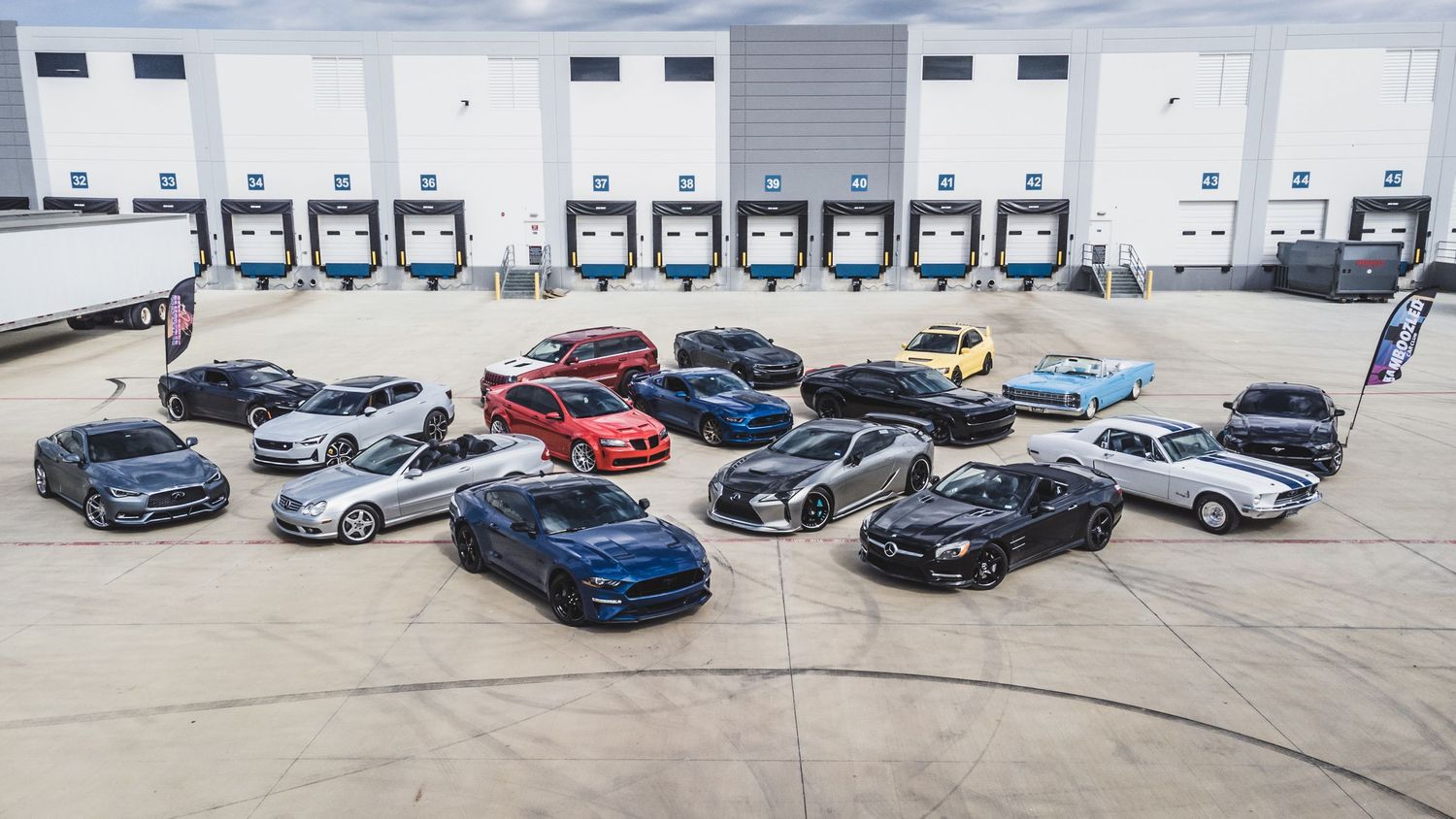 Collection of sports cars arranged in a semi-circle in front of loading docks at a warehouse.