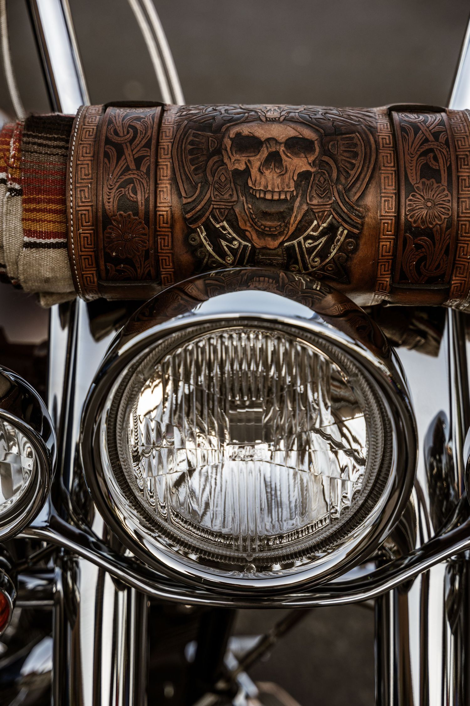 Custom leather motorcycle handlebar cover with ornate skull design above chrome headlight.