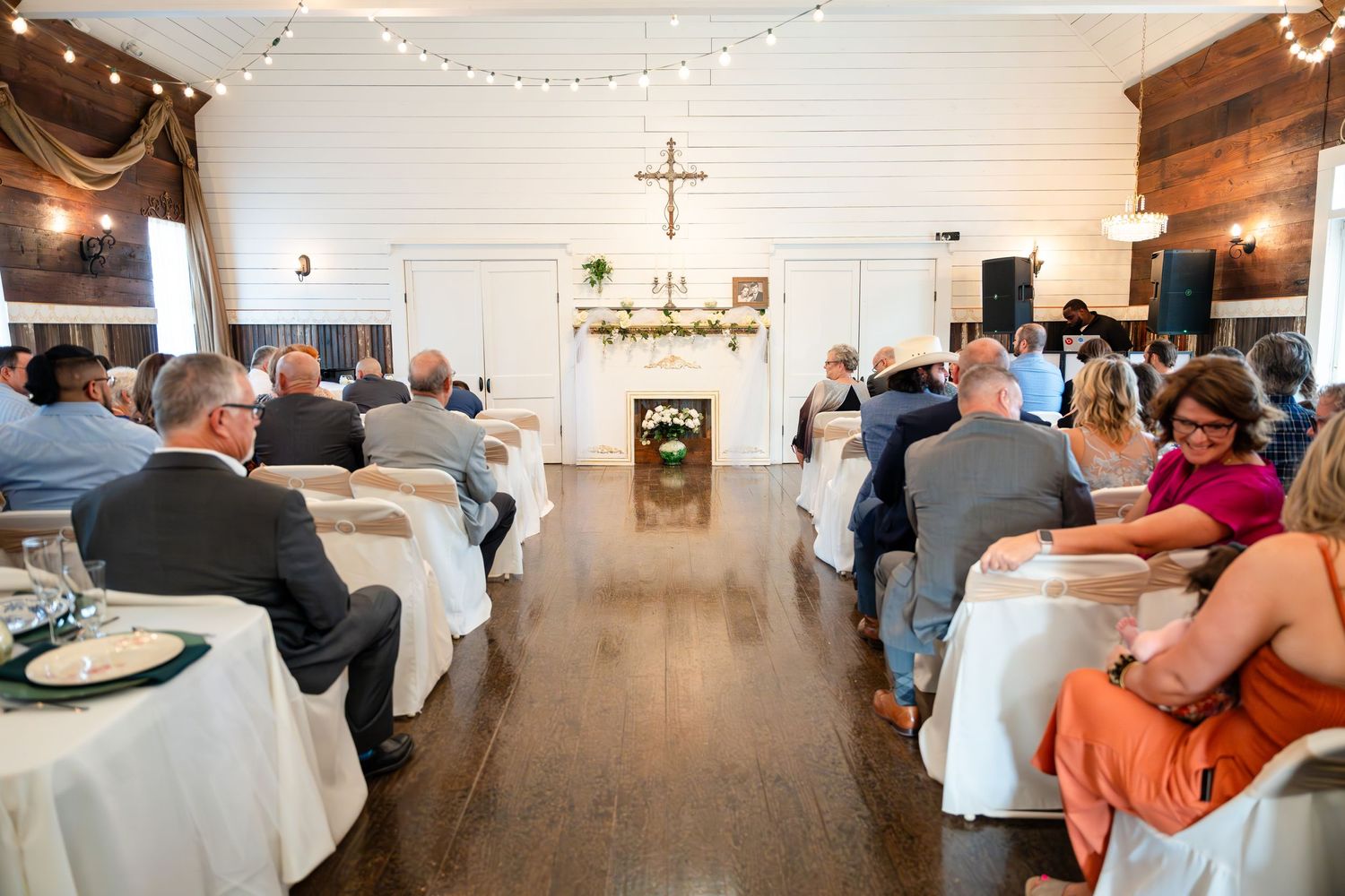 Wedding ceremony in a rustic white chapel with wooden floors and string lights overhead.
