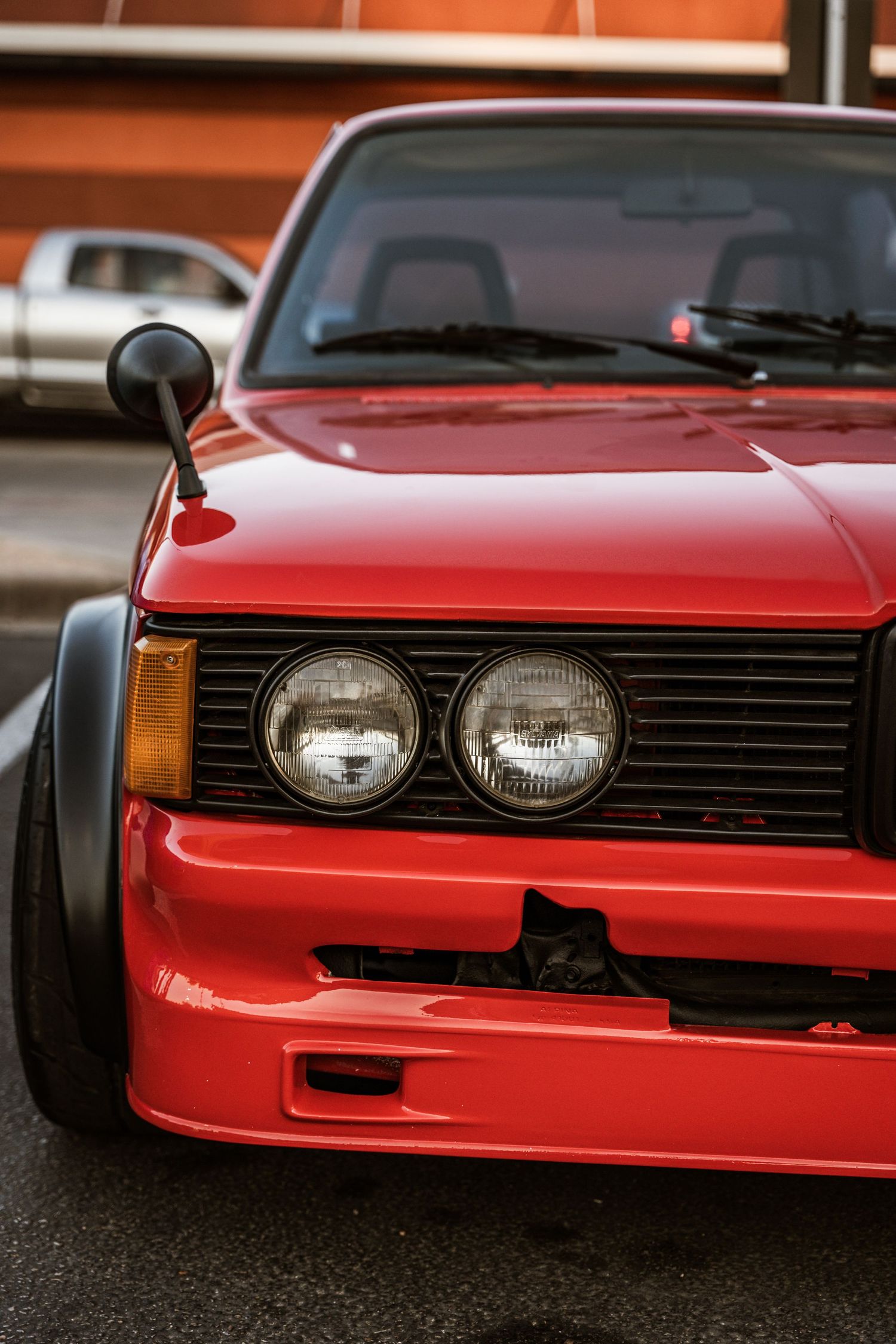 Close-up view of red vintage car front end showing double headlights and modified body kit.