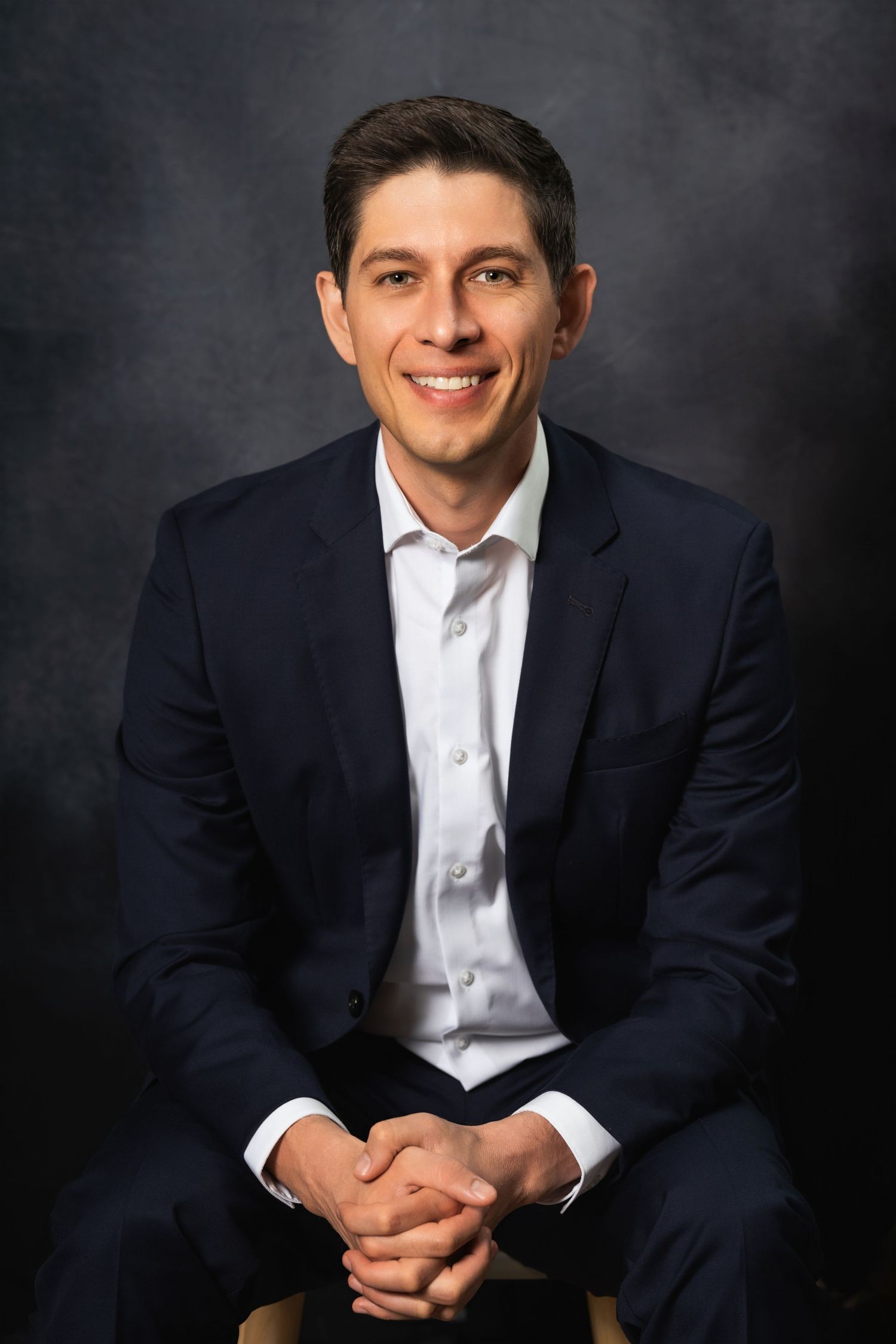 Professional portrait of a seated person in business attire with clasped hands.