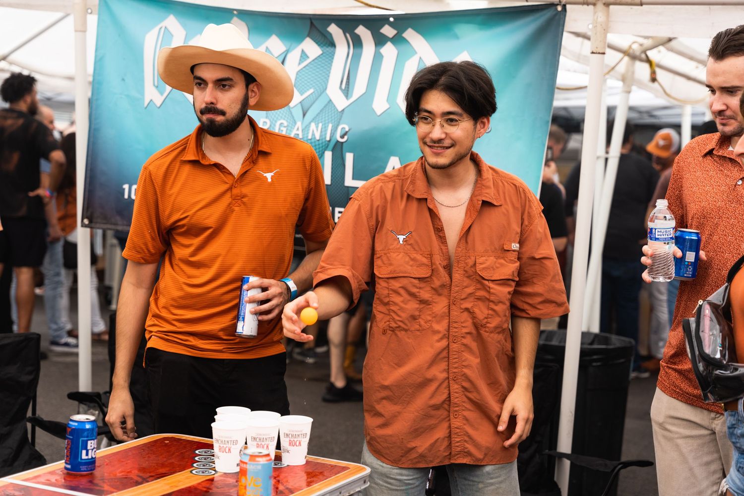 Two people in orange shirts enjoying drinks under a blue tent banner at an outdoor event.