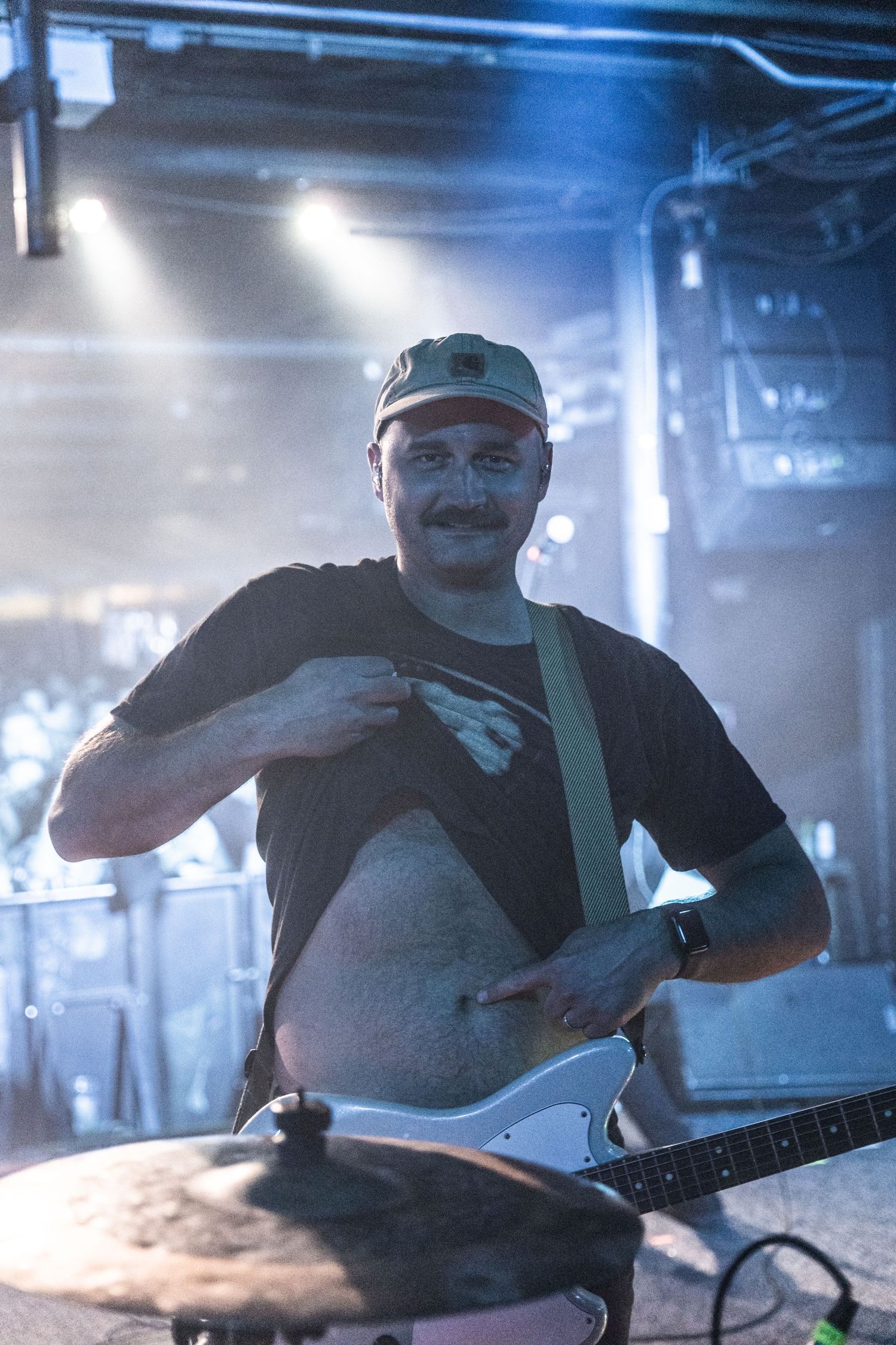 Performer lifts shirt while playing guitar during concert with blue stage lighting.