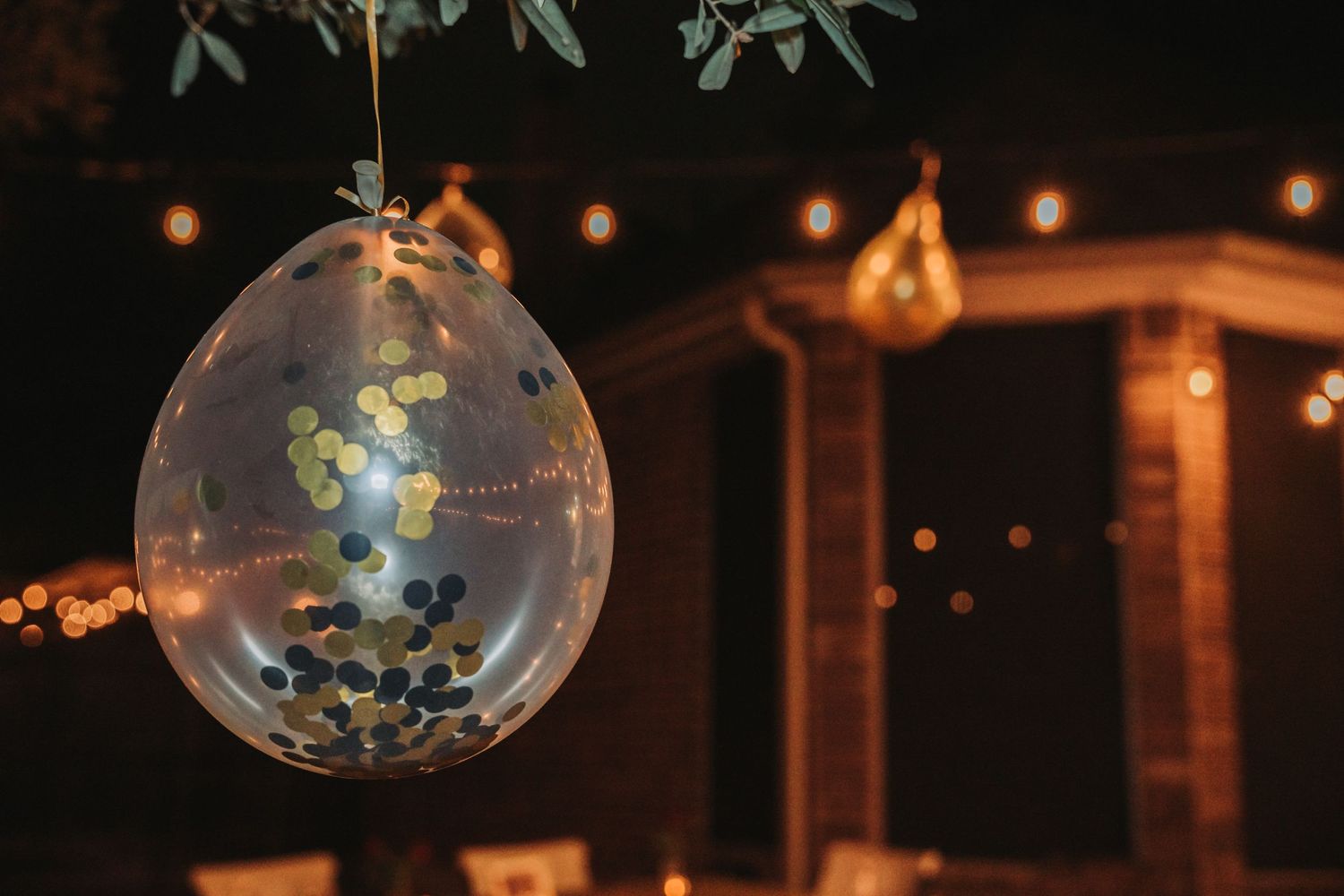 Clear balloon filled with blue confetti hangs from foliage with glowing lights in background.