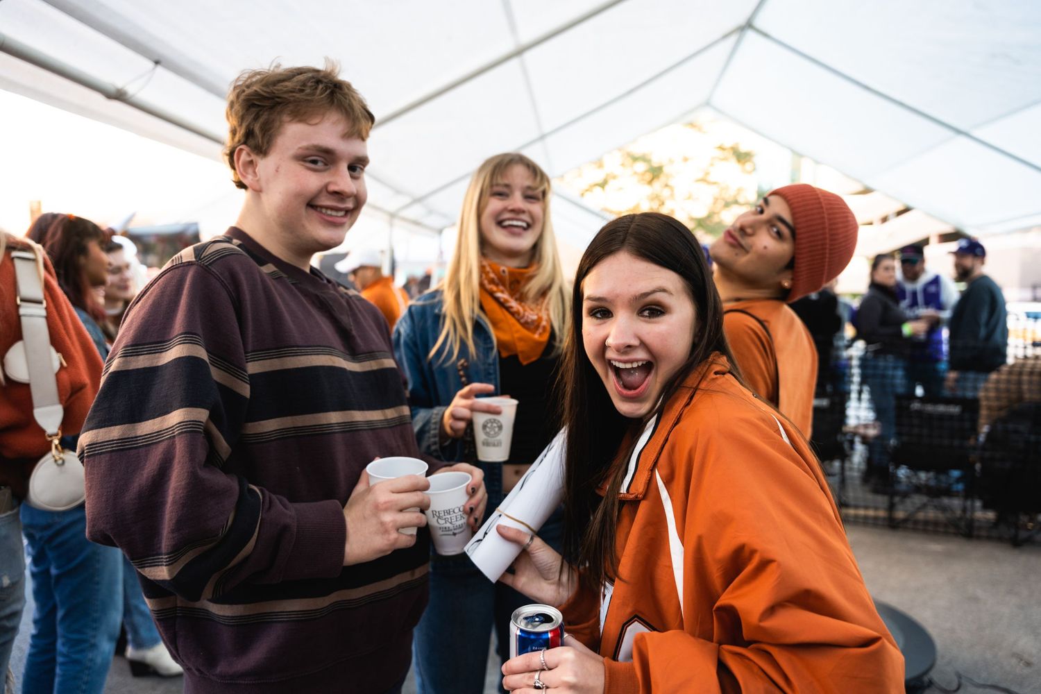 Friends enjoying drinks and laughing together at an outdoor event under a white tent.