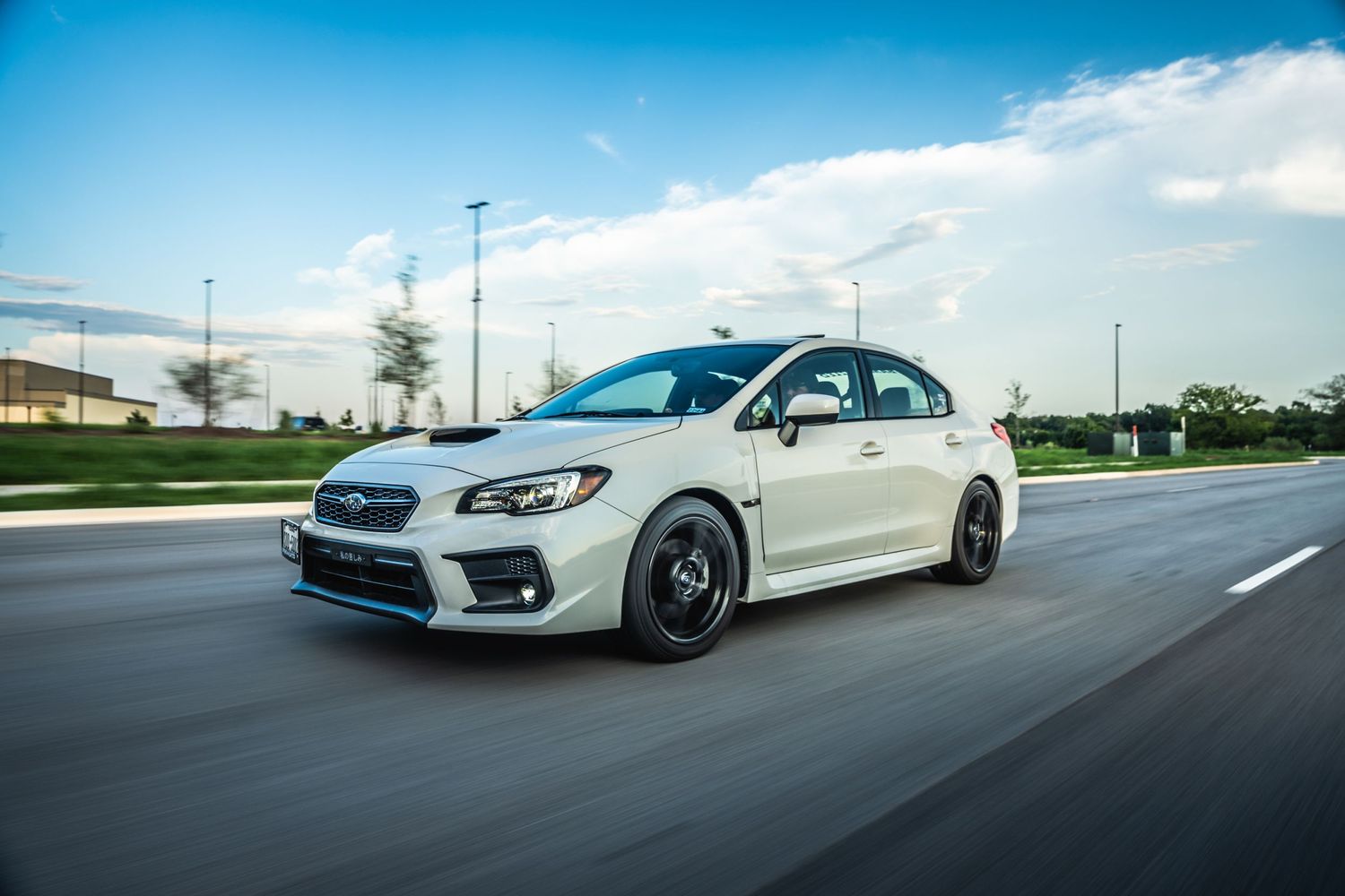 White Subaru WRX sedan with black wheels driving on a highway under blue skies.