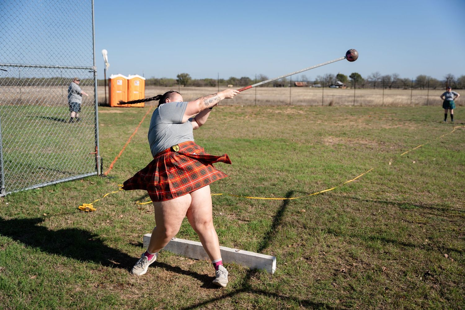 Person in tartan kilt throwing hammer at Highland Games competition on grassy field outdoors.