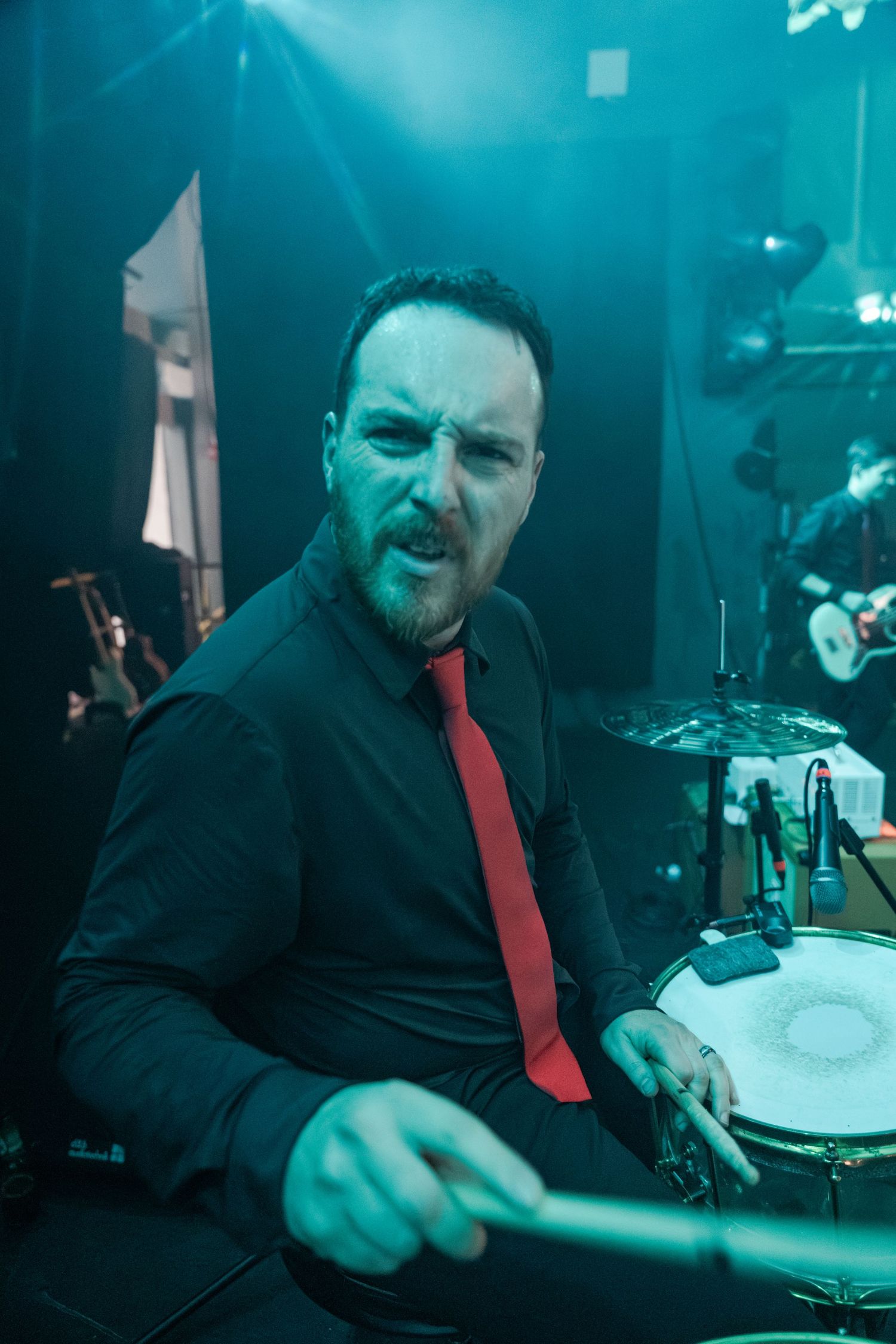 Male drummer in black shirt and red tie playing drums on stage with teal lighting during live performance.