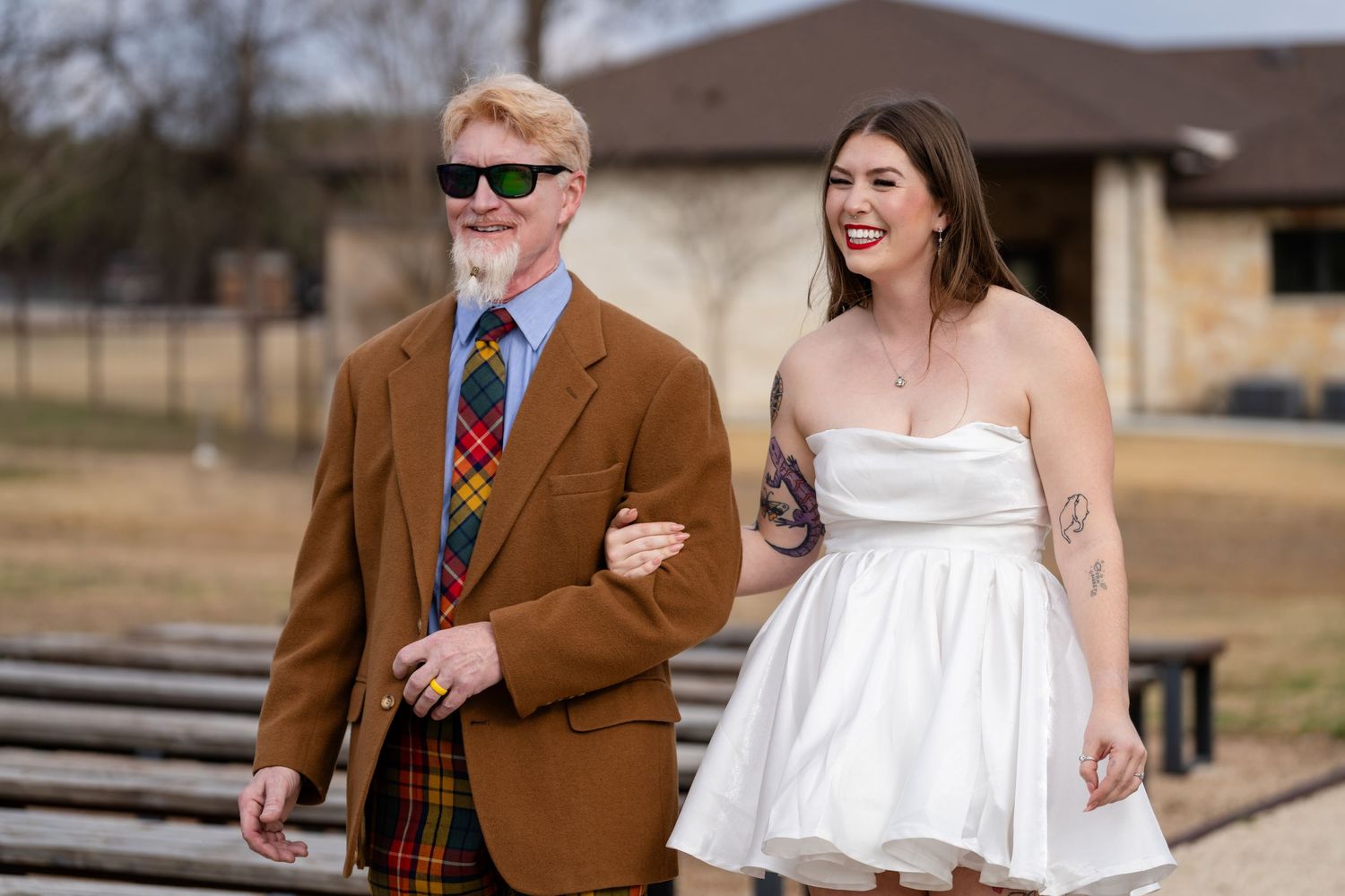 Couple walking together outdoors, man in brown plaid suit with colorful tie, woman in white strapless dress with tattoos.