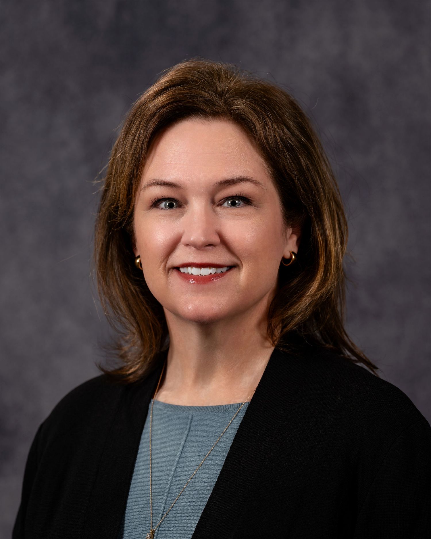 Professional headshot of a smiling woman with brown hair wearing a black blazer and light blue top against a gray background.