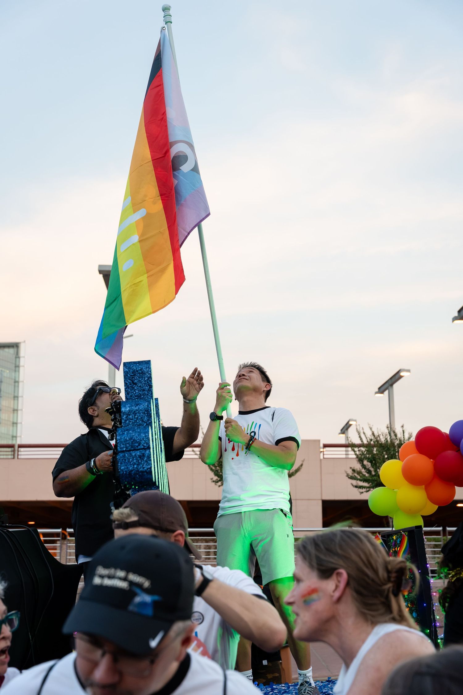 Person proudly raising a rainbow Pride flag at an outdoor LGBTQ+ Pride celebration with colorful balloons and crowd.