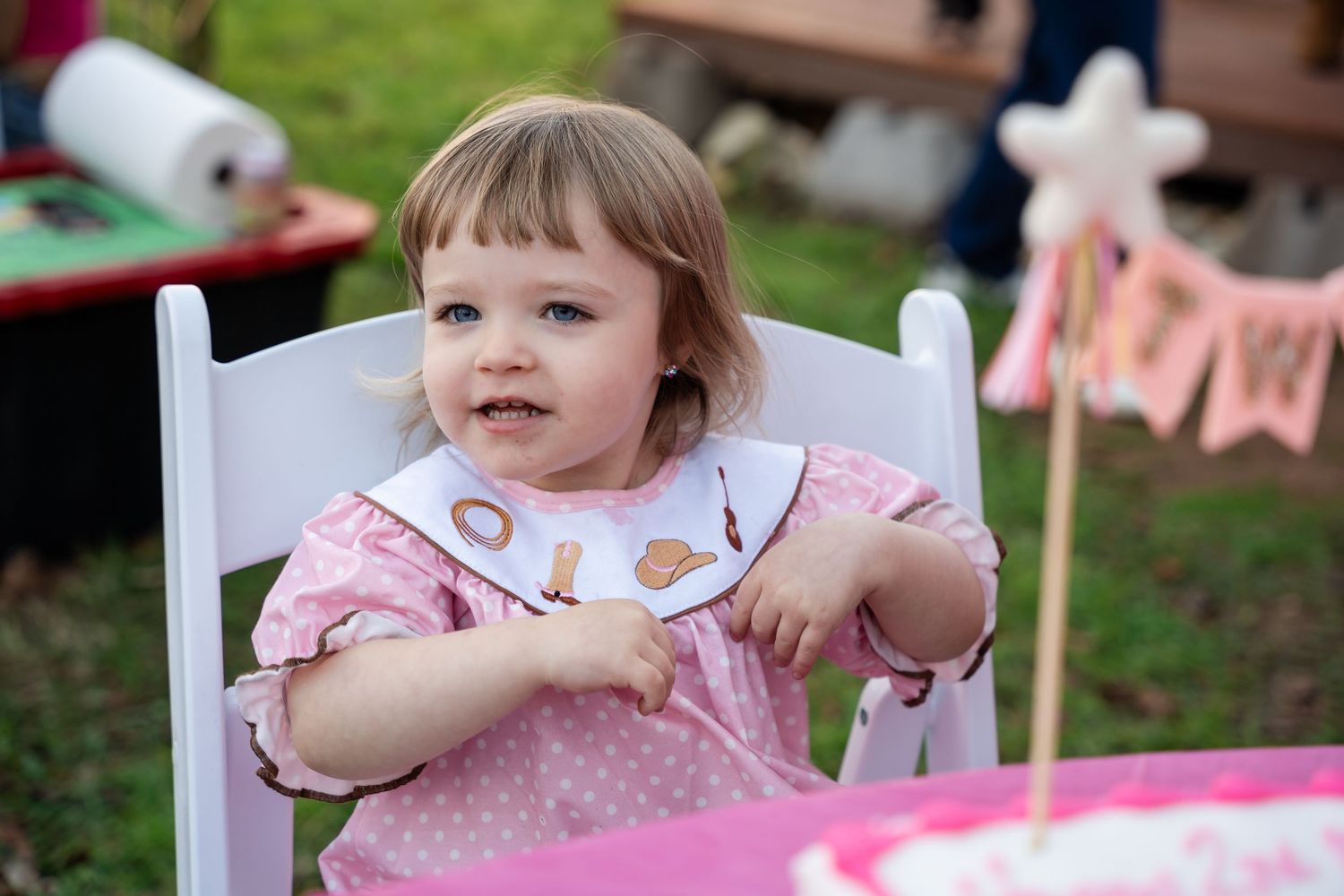 A child sits in a white chair at a pink-themed party with decorative banners in the background.