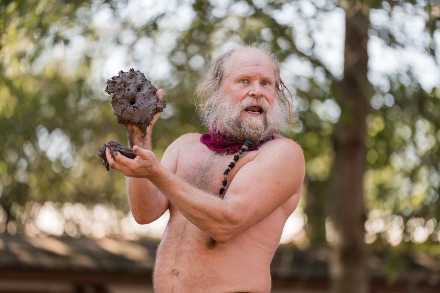 Shirtless person with long white hair and beard juggling rocks outdoors in a forest setting.