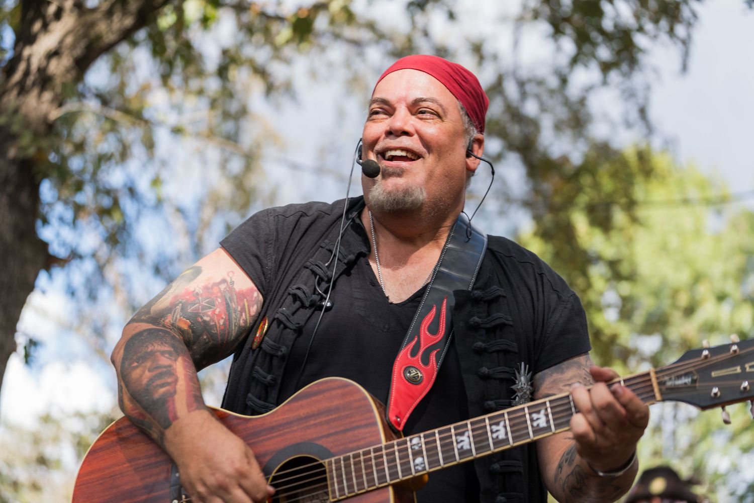 Musician playing acoustic guitar and singing outdoors with trees in background.